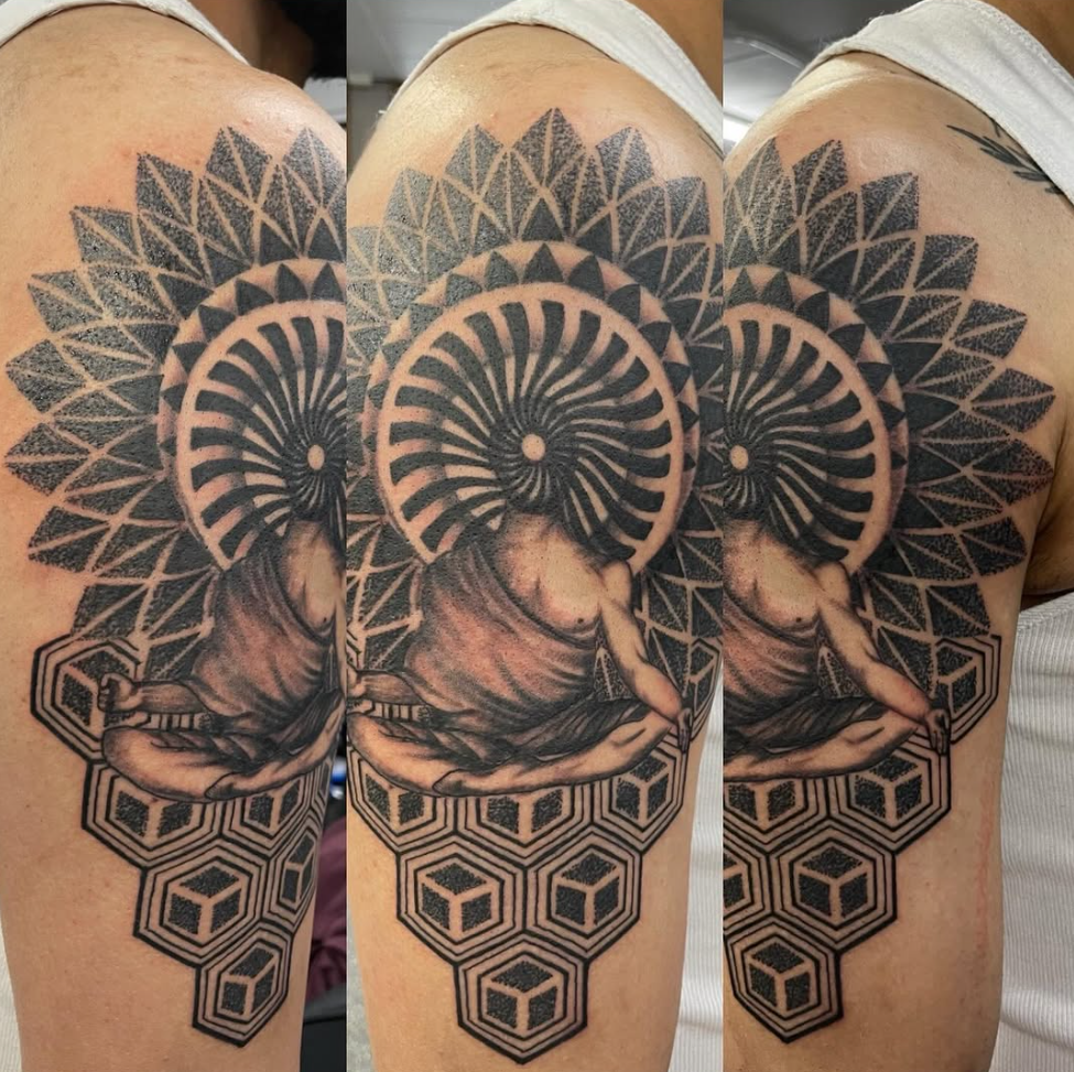 A detailed black and gray tattoo on a person's shoulder and upper arm, featuring a person in a seated meditative pose with a spiral shell or pattern above their head, surrounded by geometric and hexagonal patterns.