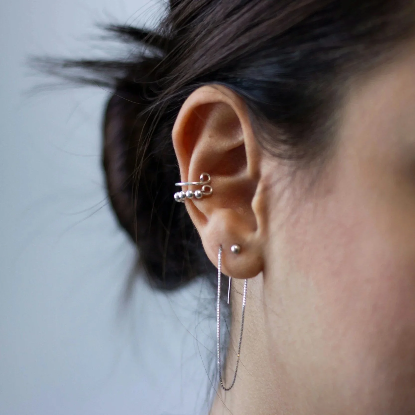 Close-up of a person's ear with multiple silver earrings and a necklace, with dark hair and a plain background.