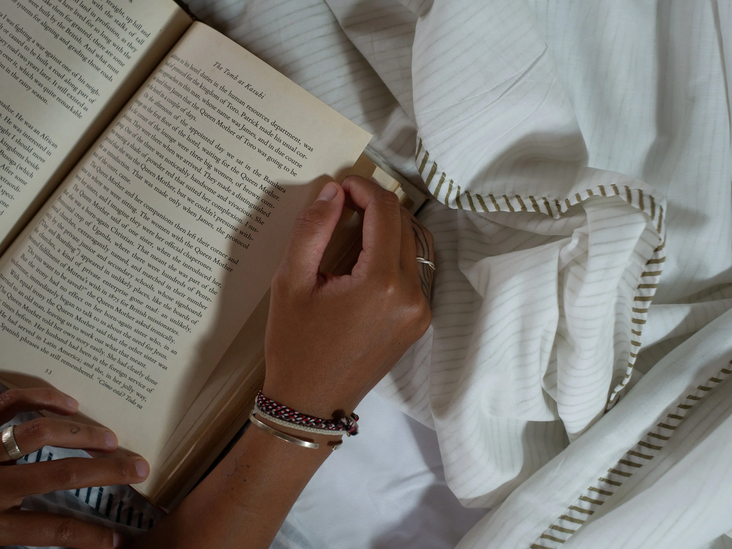 A oeorson comfortably reading a book while lying on a bed covered with white Cedar dcanter muslin blanket