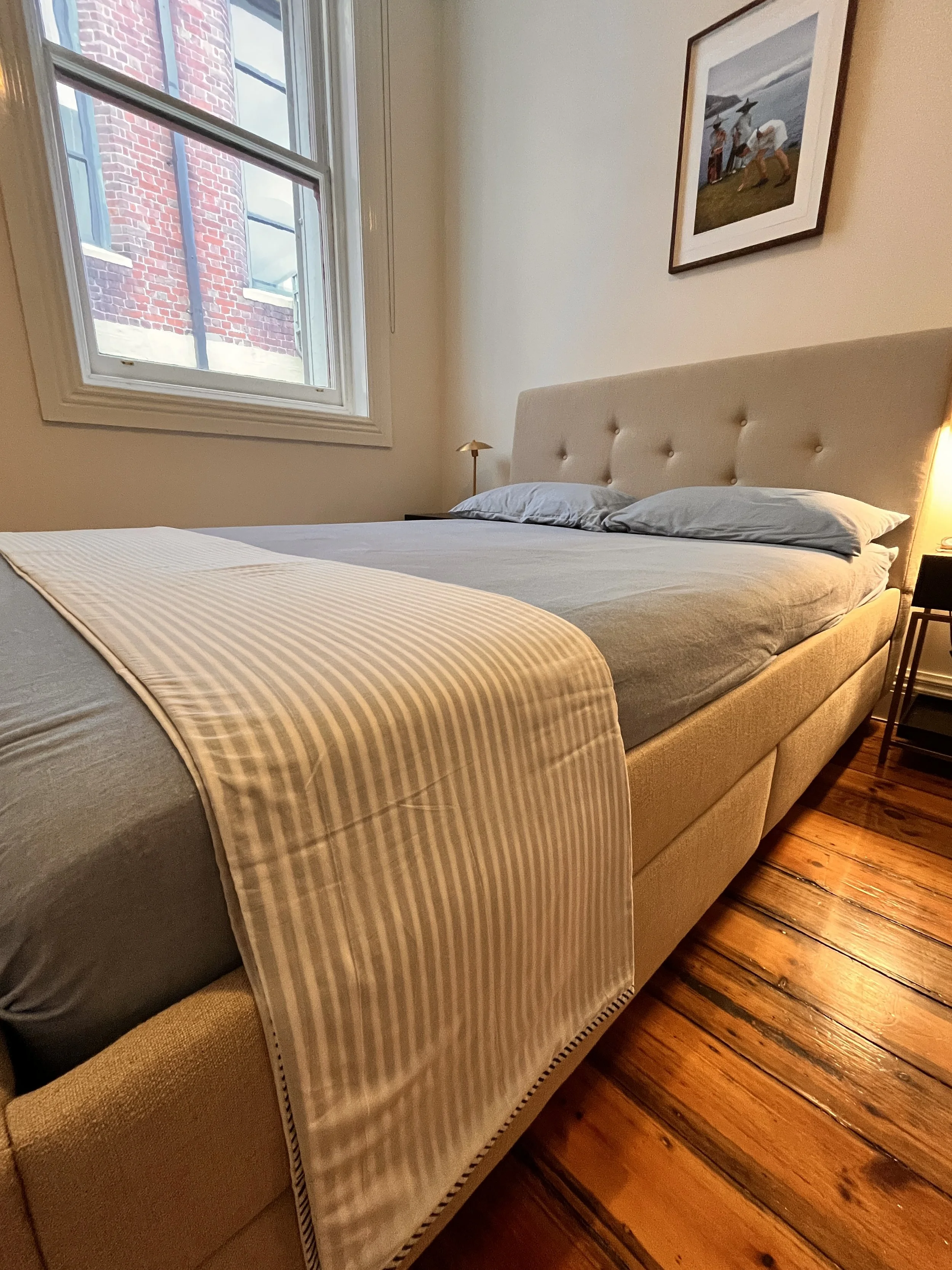 A bedroom with a window showing a brick building outside. The bed has a beige headboard, a gray striped blanket, and gray pillows. There is a small gold-colored lamp and a framed picture of birds on a beach hanging on the wall.