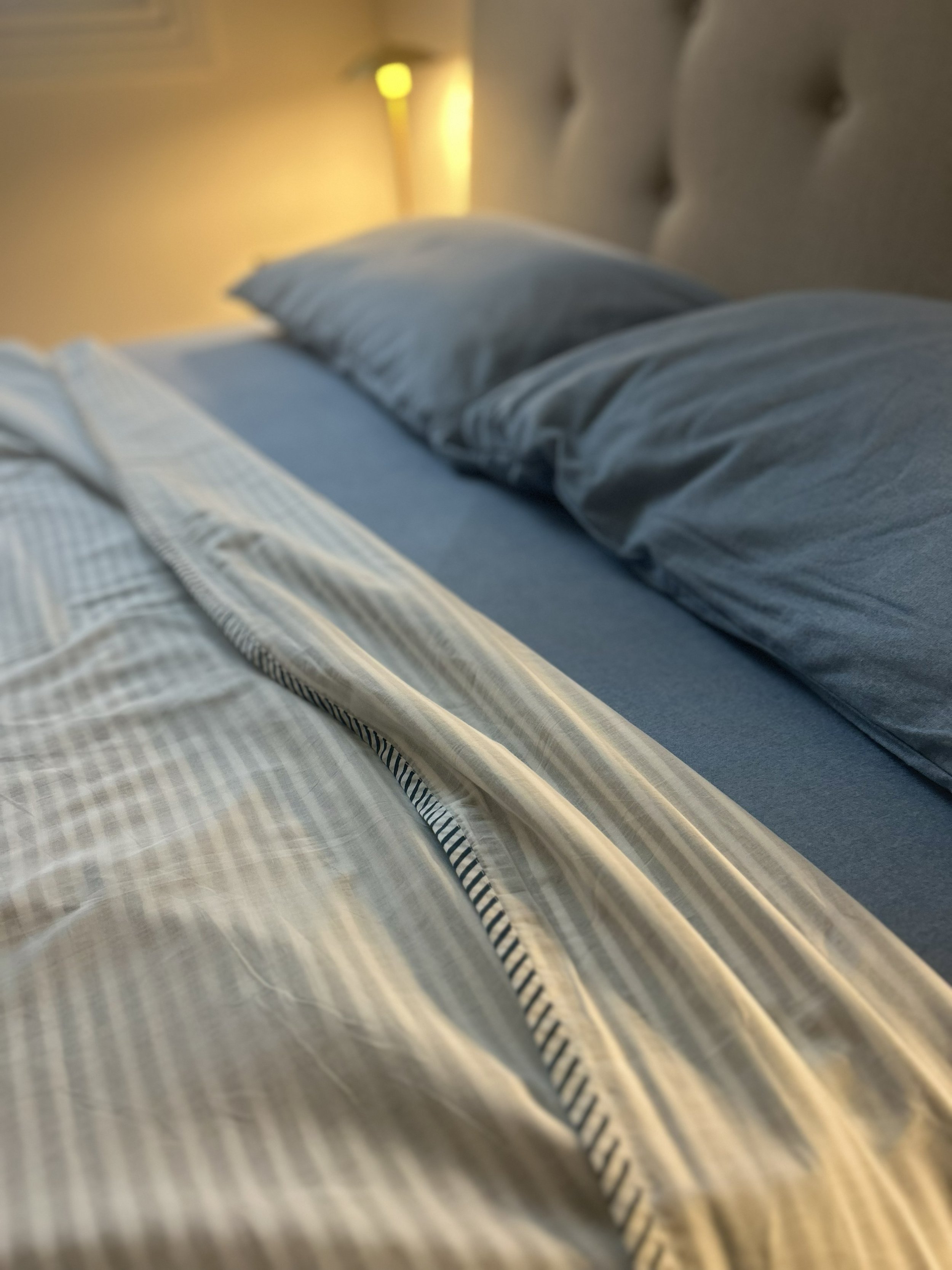 A bed with a gray headboard, blue sheets, and striped pillows in a bedroom with a bedside lamp.