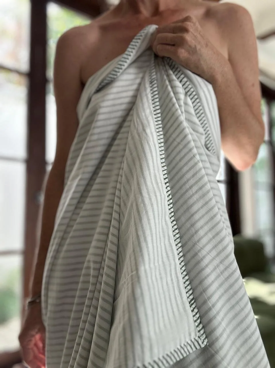 An elderly person holding a striped cloth or towel against their chest, wearing no shirt, with a background of a room with large windows and natural light.