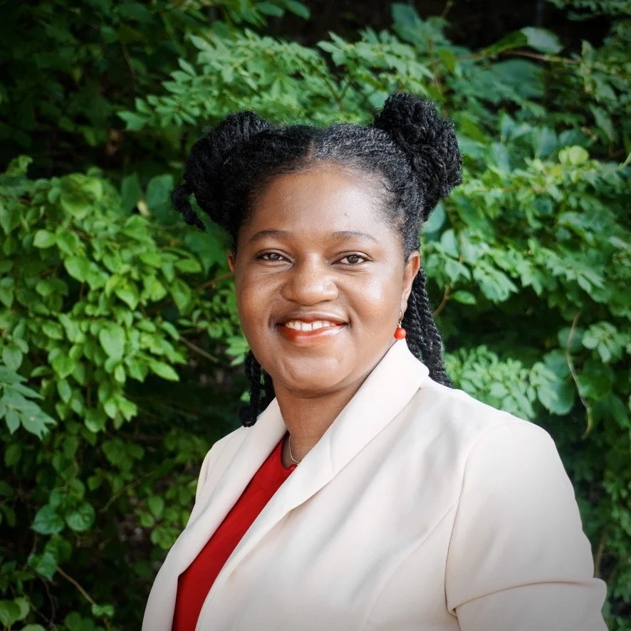 A woman with braided hair styled in two buns, wearing a white blazer over a red top, and earrings, standing outdoors in front of green leafy bushes.