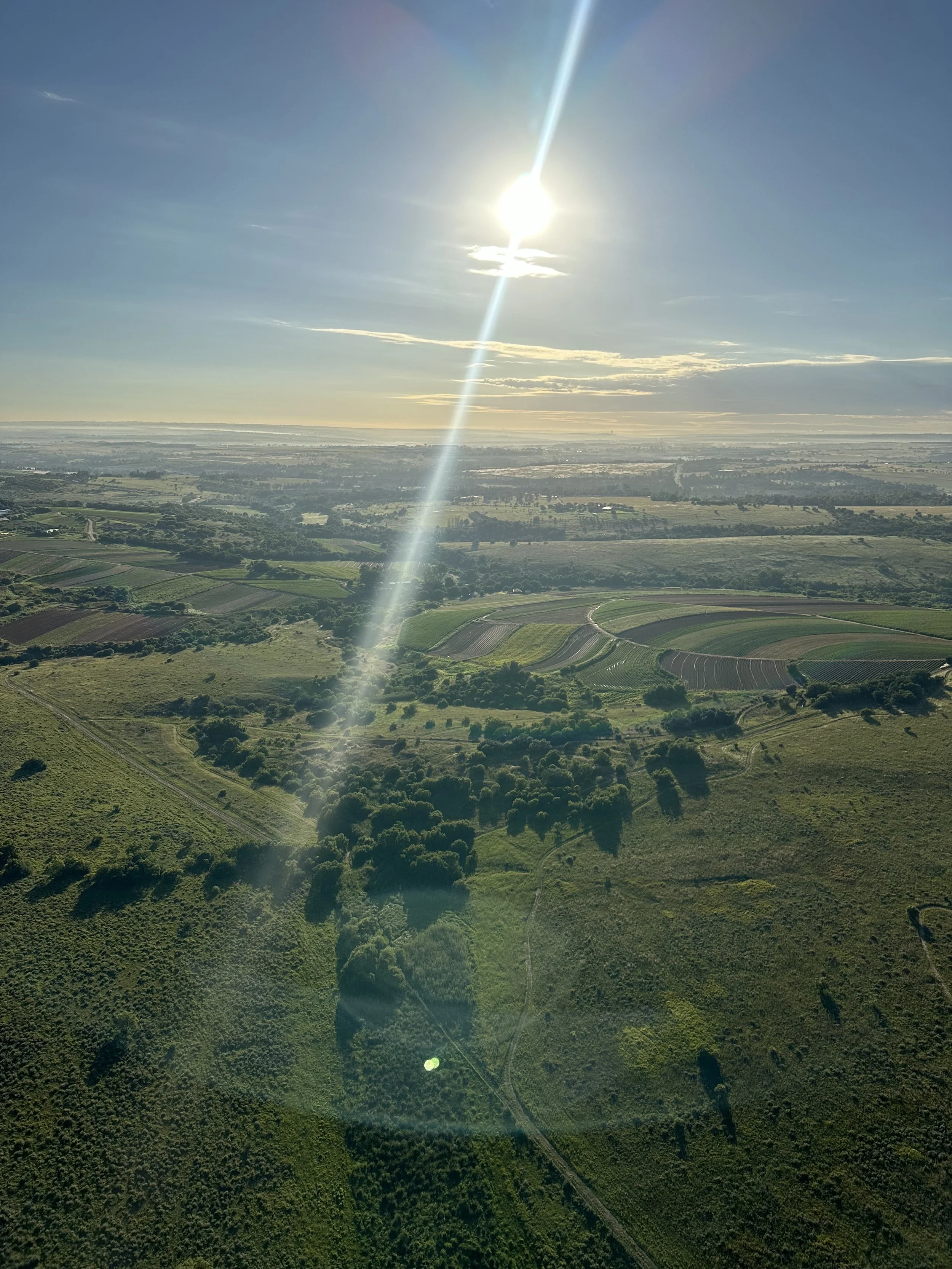 Aerial view of a vast landscape with green fields, patches of trees, and winding roads, under a bright sky with the sun shining and some clouds.