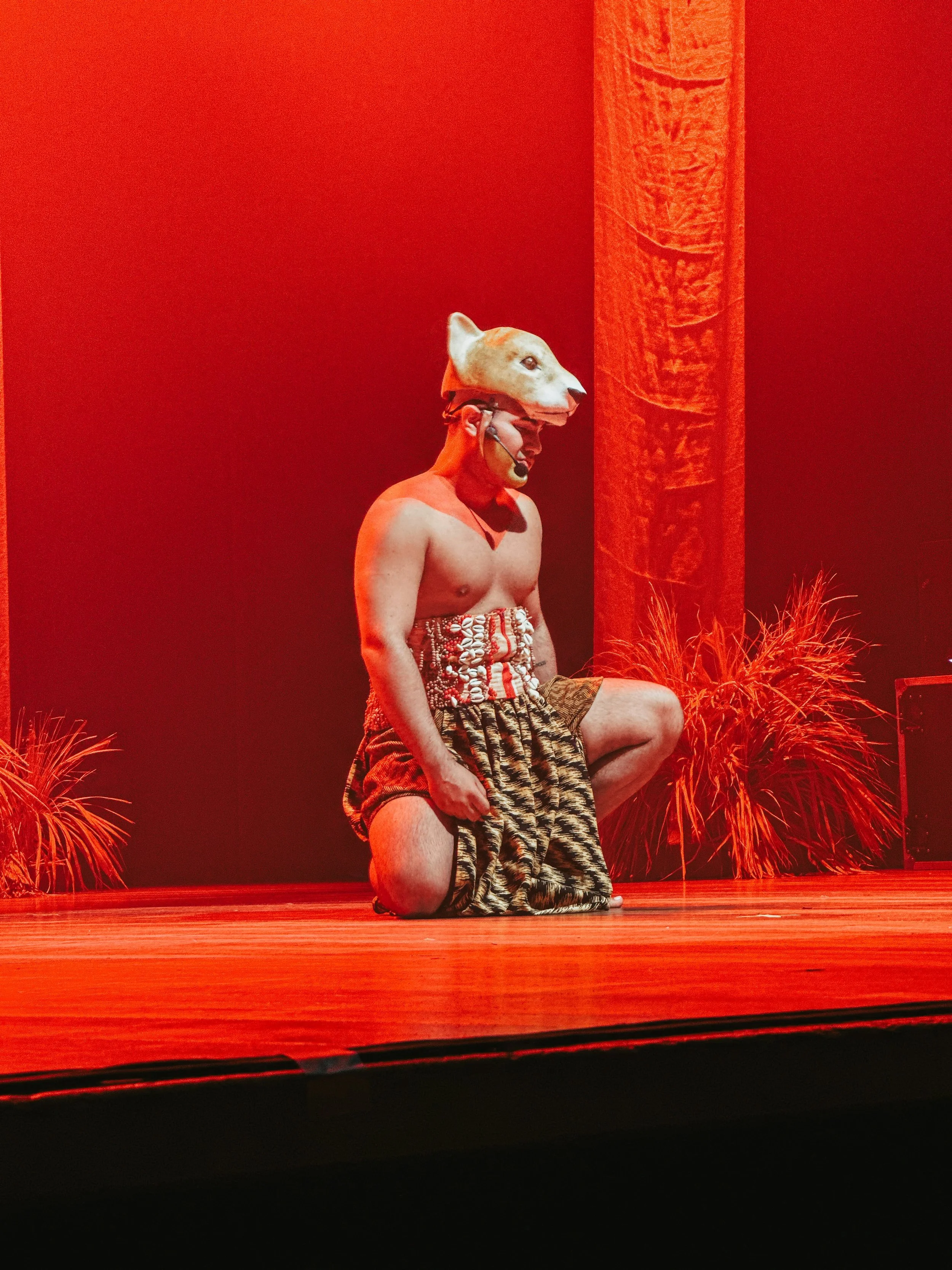 Performer wearing a wolf mask with a human body, dressed in traditional tribal attire, kneeling on one knee on a wooden stage with red lighting and tribal decor in the background.