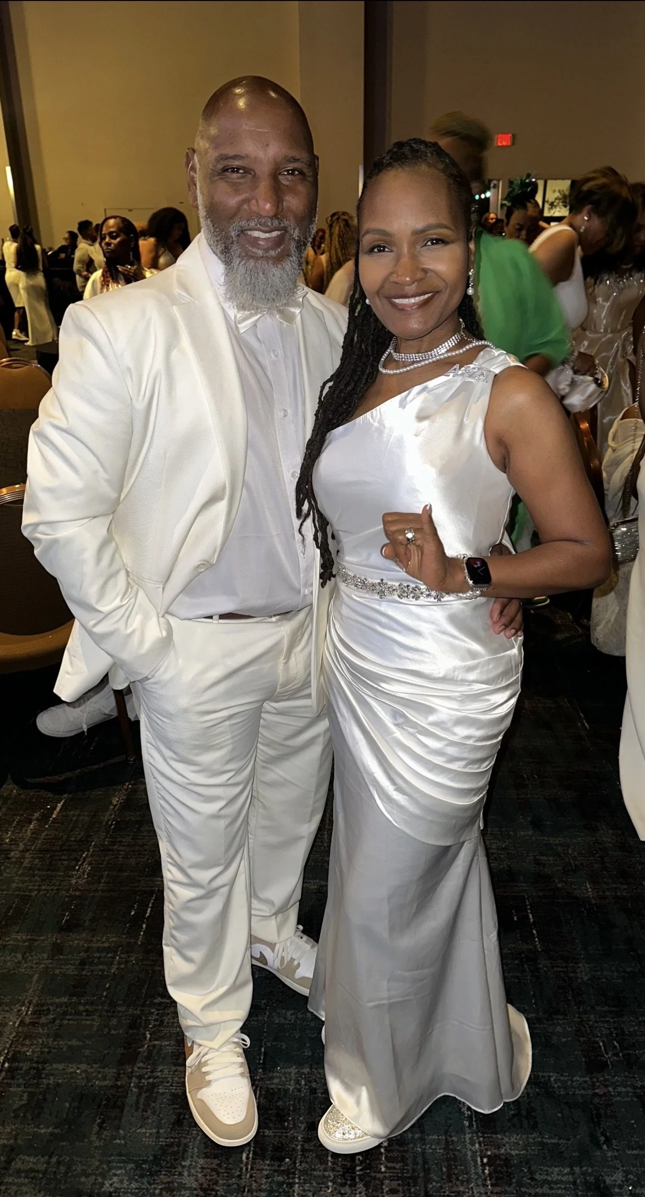 A man and woman dressed in white at a formal event, standing together and smiling, with other people in the background.