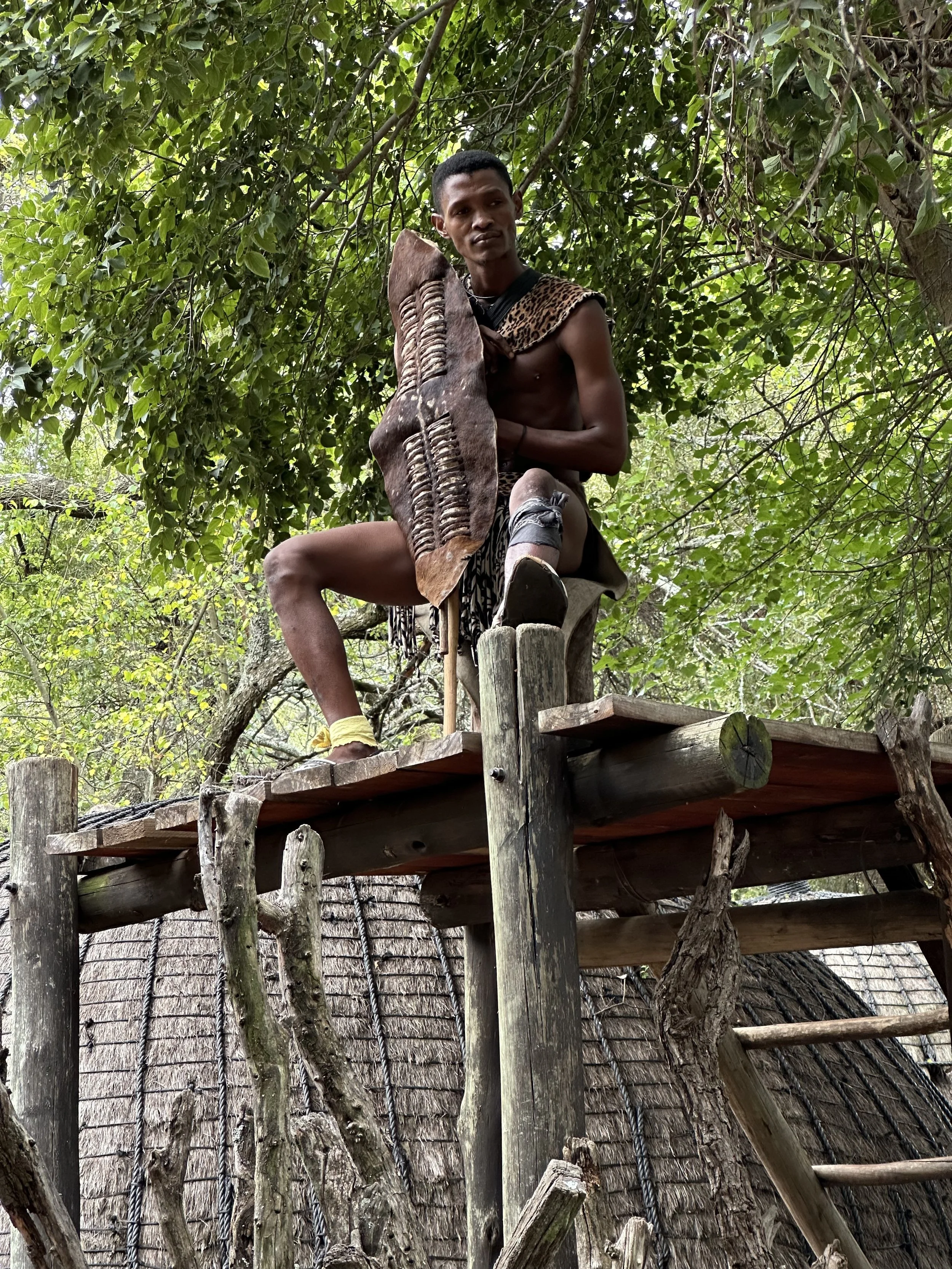 A person with dark skin, short black hair, sitting on a wooden platform high in a tree, surrounded by green leaves, holding a wooden carving, wearing animal print clothing and a leg band, with a thatched roof structure below.