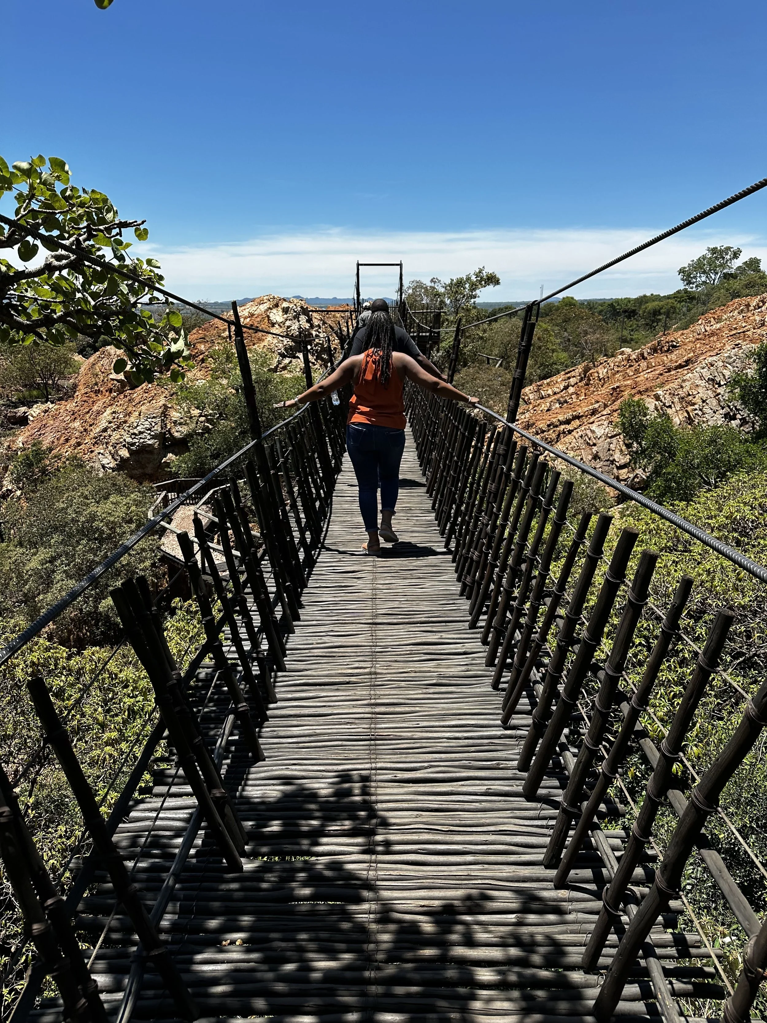 People walking on a wooden suspension bridge in a natural landscape with rocks, trees, and a blue sky.