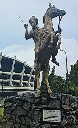 Bronze statue of a man on horseback holding a sword, mounted on a stone pedestal in an outdoor park, with a modern building and trees in the background.
