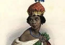 Portrait of a girl wearing a traditional crown, necklace, and a decorative dress with a bow, posing against a plain background.