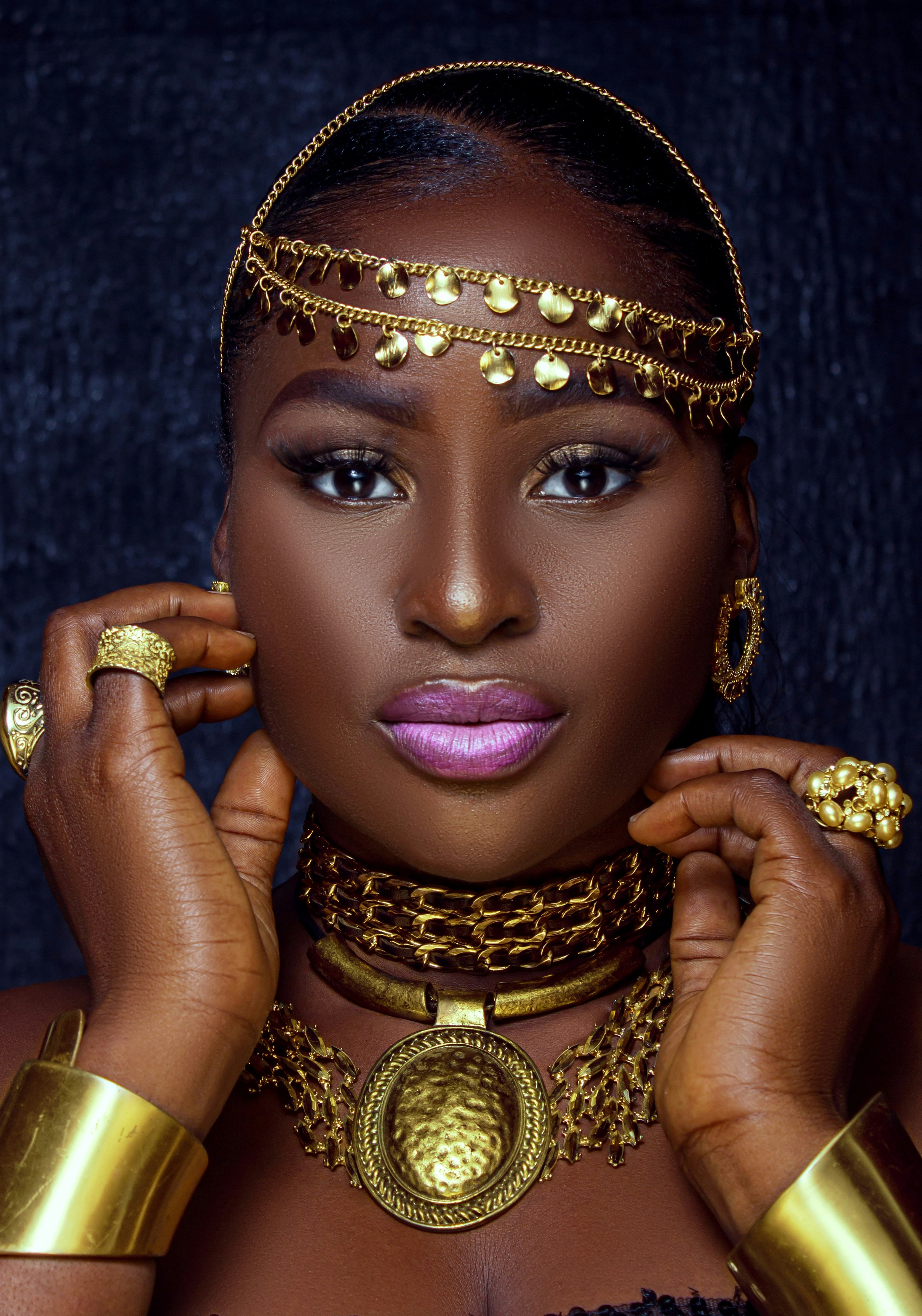A woman wearing multiple gold jewelry pieces including a layered chain headpiece, large earrings, a thick choker, a large medallion necklace, and wide bangles. She has dark skin, shiny makeup, and black hair styled close to the scalp. Her hands are touching her face, showcasing rings and bangles.