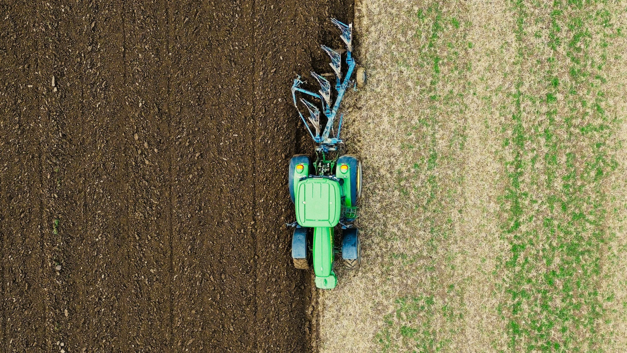 A green tractor with a blue plow attached, working on a field with one side freshly tilled soil and the other side with grass and small plants, seen from above.