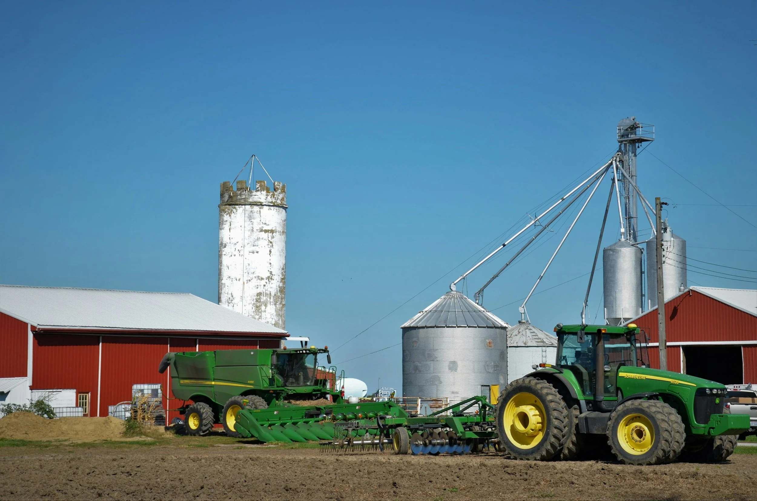 Farm scene with green tractors, metal silos, a red barn, and farm equipment under a clear blue sky.