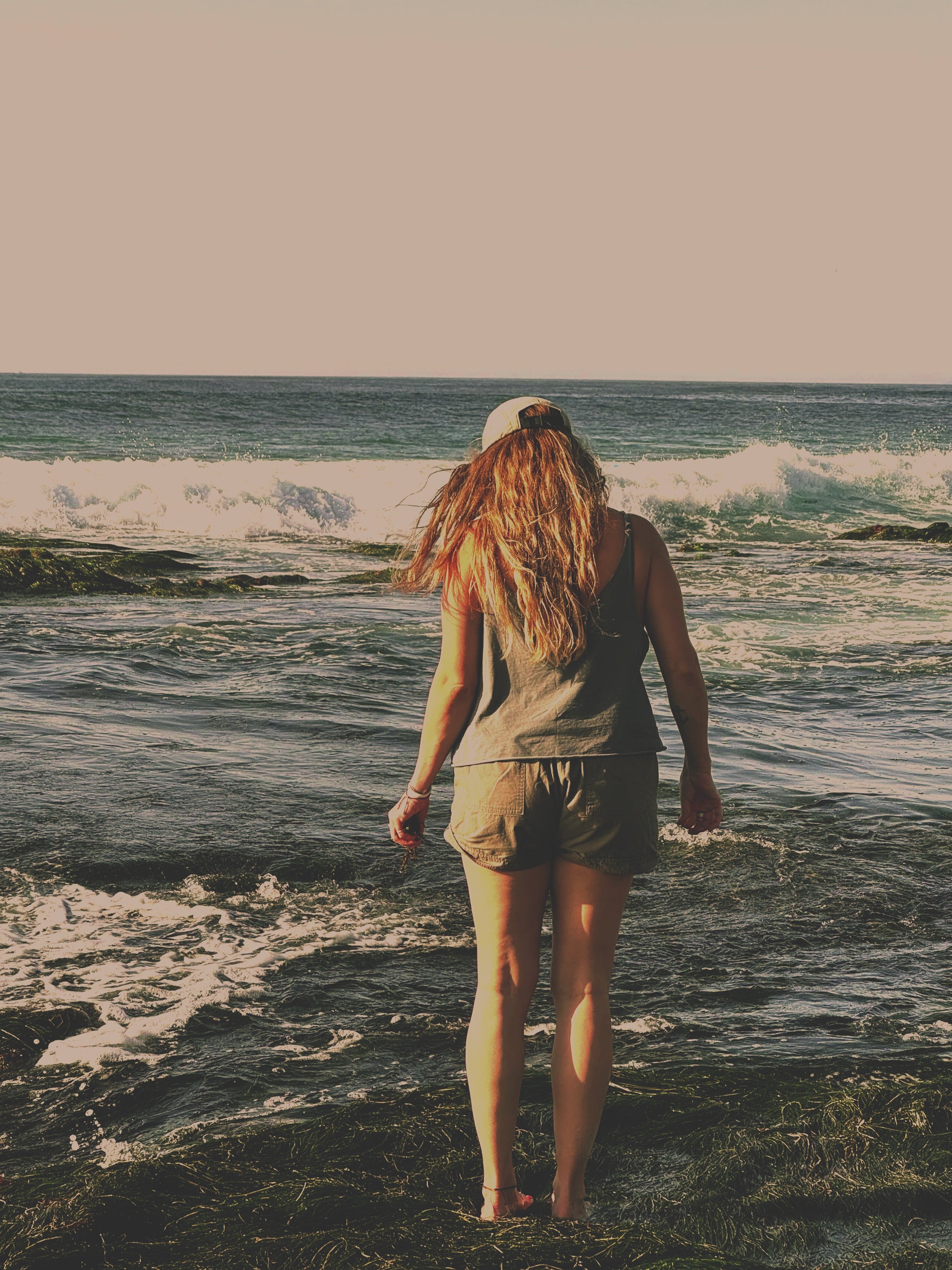 A person with long brown hair wearing a gray tank top, khaki shorts, and a white cap stands at the edge of the ocean, facing away, with waves crashing near their feet during sunset or sunrise.