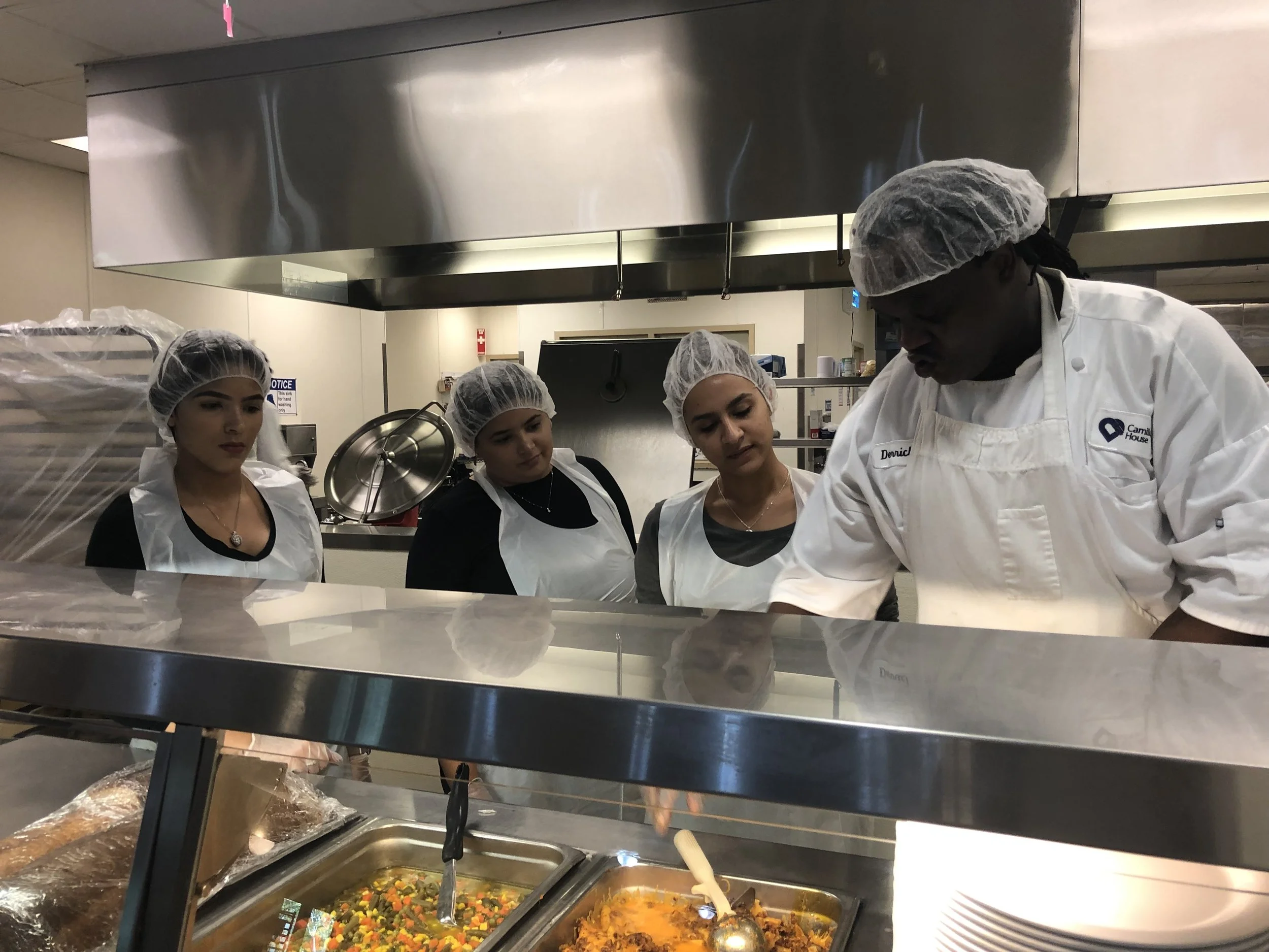 A group of chefs wearing hairnets and aprons working behind a food serving counter with dishes of vegetables and cooked meats. The scene appears to be in a commercial kitchen or cafeteria.