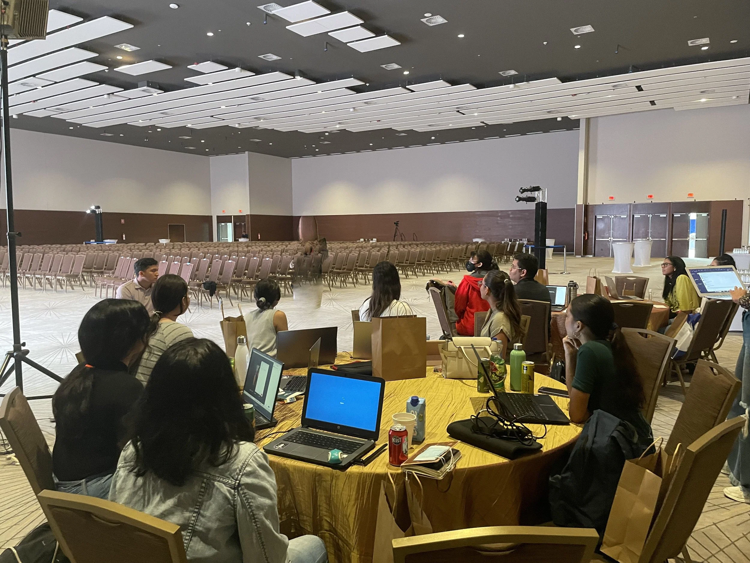 A large conference or seminar room with many empty chairs arranged in rows and a few people seated at a round table in the foreground, working on laptops. The room has high ceilings with modern light fixtures and a neutral color scheme, with some people wearing masks.