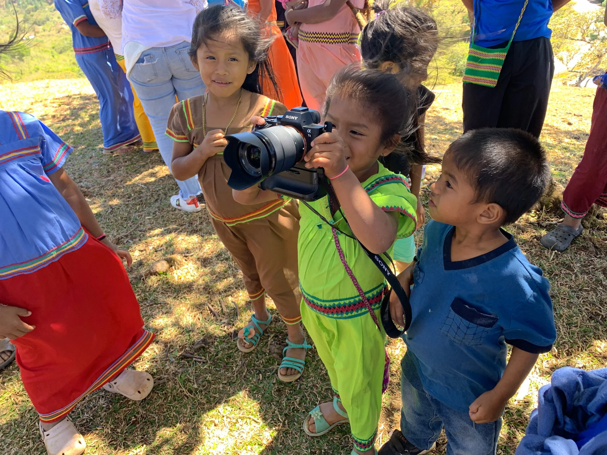 Young children dressed in colorful traditional clothing gathered outdoors, some are smiling and one girl is holding a camera.