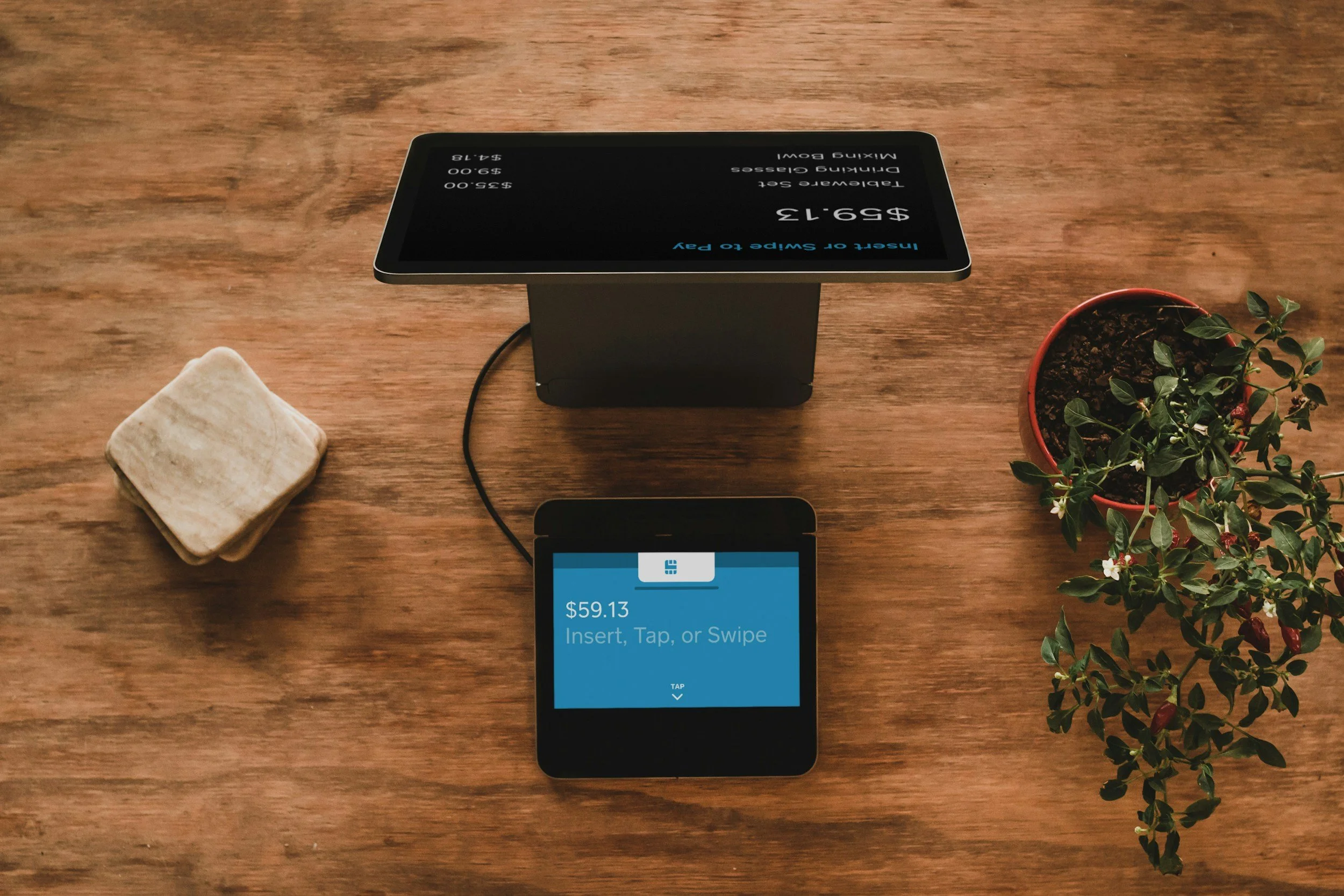 A wooden table with a small square tan stone object on the left, a black digital payment device with a stand in the center, and a potted green plant with small white flowers on the right. The digital device displays a prompt about a payment of $59.13.