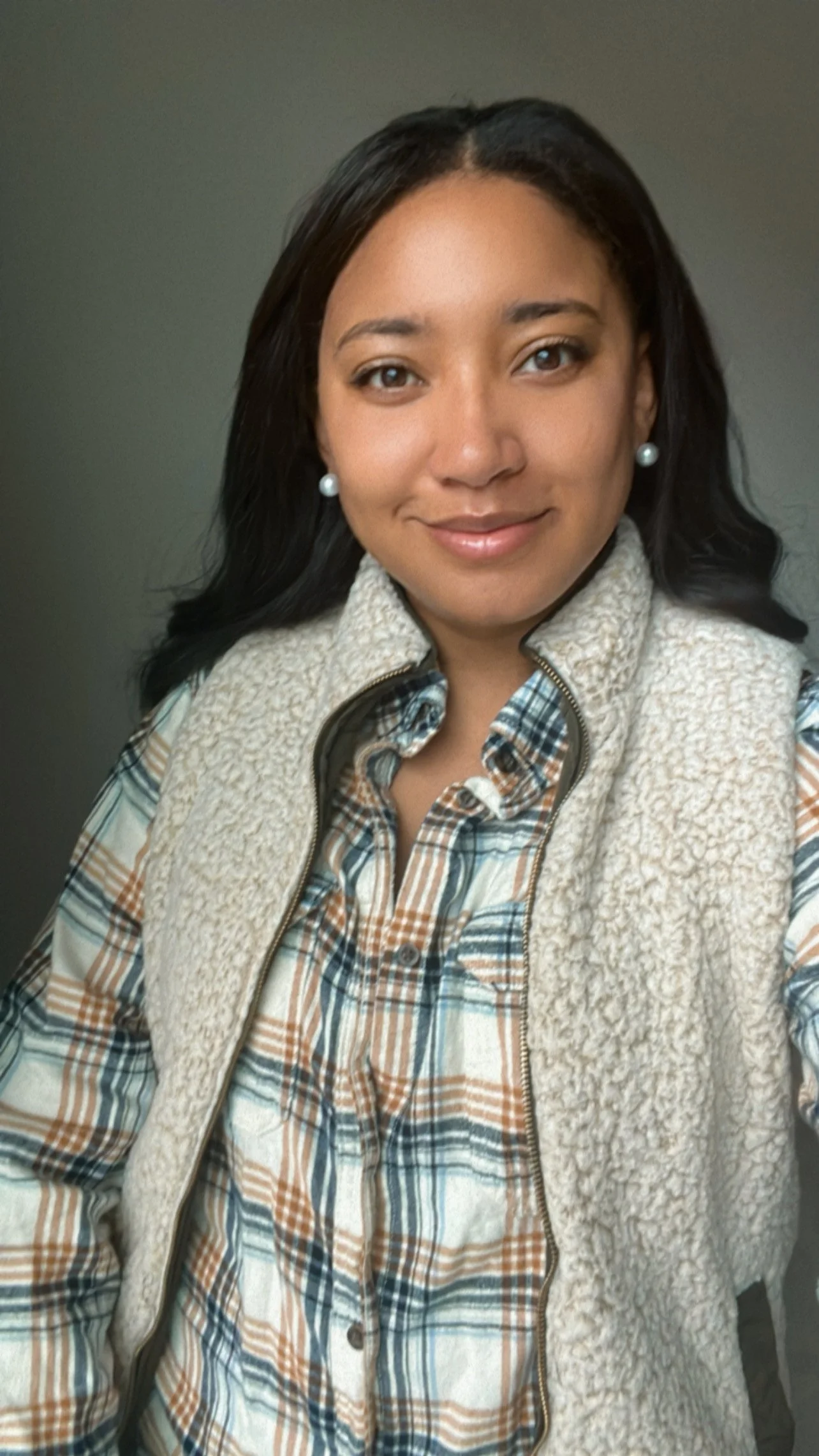 A woman with medium skin tone, black hair, and pearl earrings smiling at the camera, wearing a plaid shirt and a beige sherpa vest, against a neutral background.