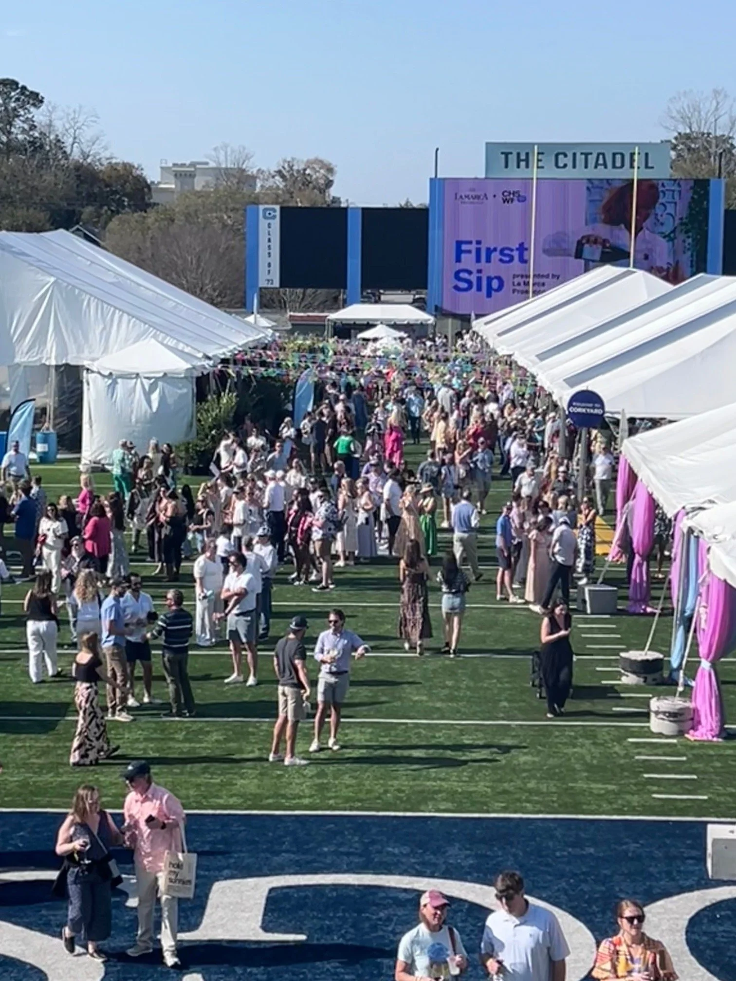 A large outdoor event with many people walking and gathering on a field, surrounded by tents and booths. There is an elevated stage or screen with banners, including one that says 'First Sip' and others promoting local brands. The area appears lively and festive.