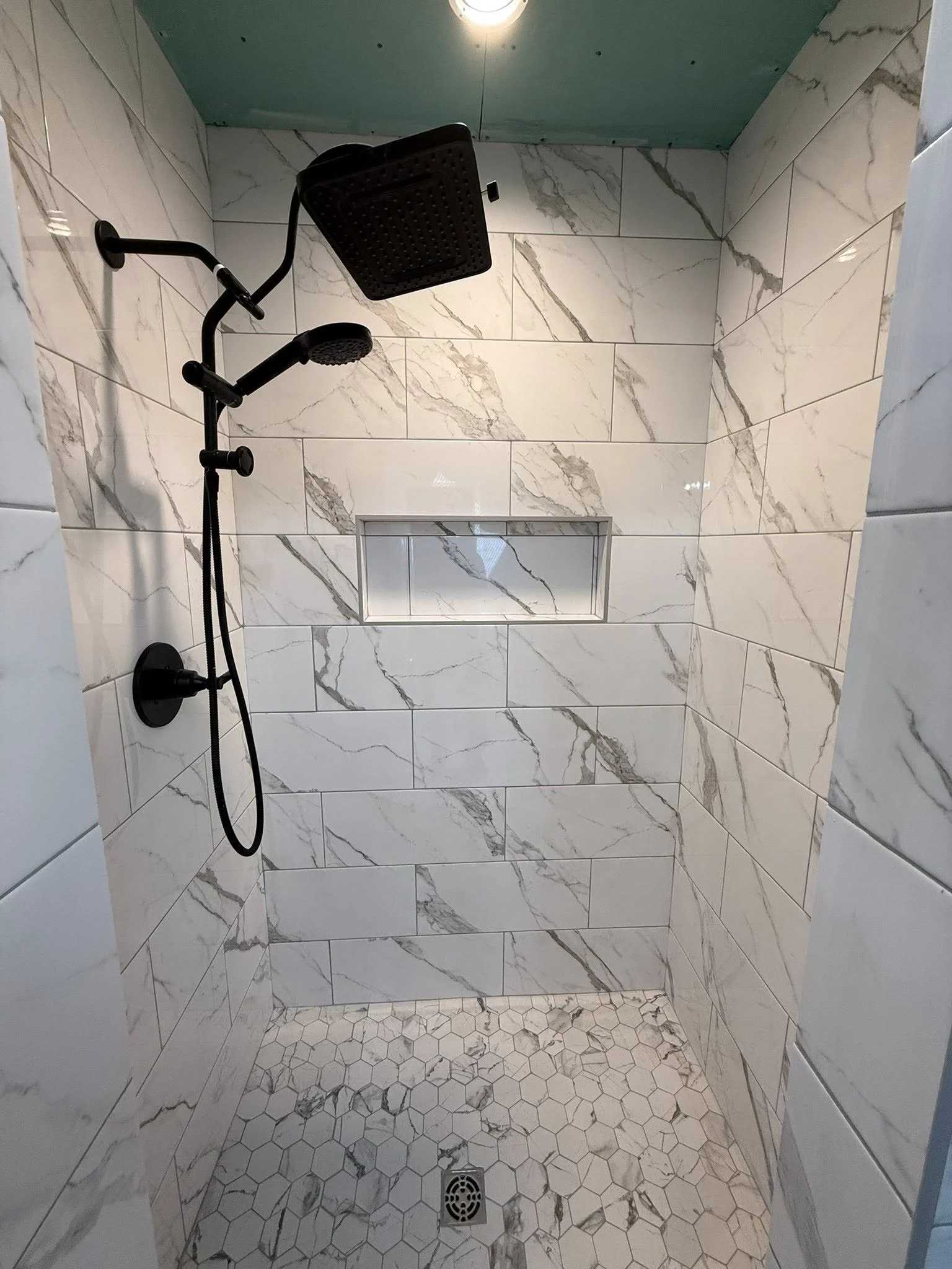 A shower with white marble walls and hexagonal marble floor tiles. It has a black showerhead and a built-in rectangular niche in the wall for storage.