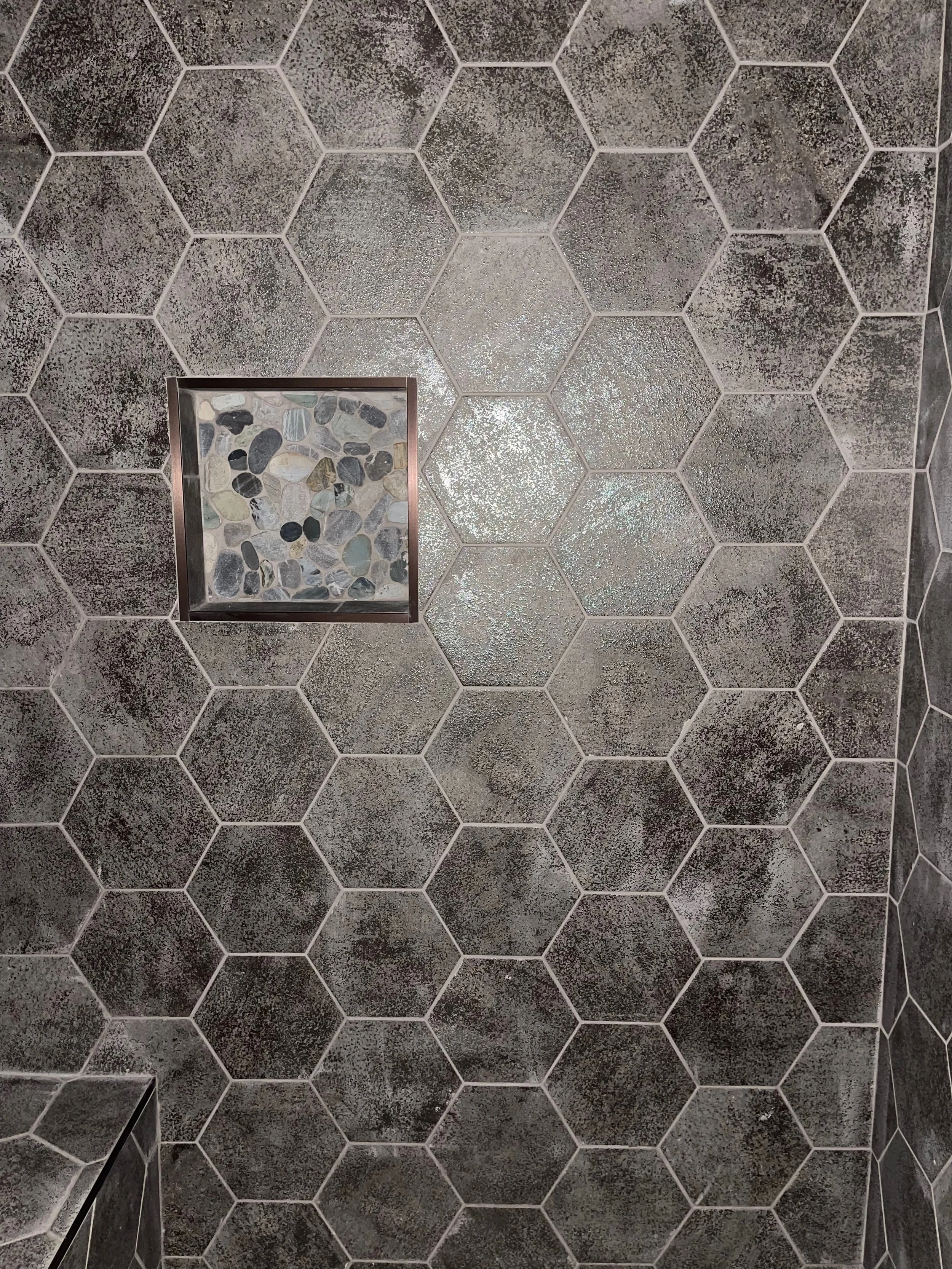 A tiled bathroom shower wall with large hexagon tiles in shades of gray and black, featuring a small built-in niche with a glass shelf and decorative pebbles.