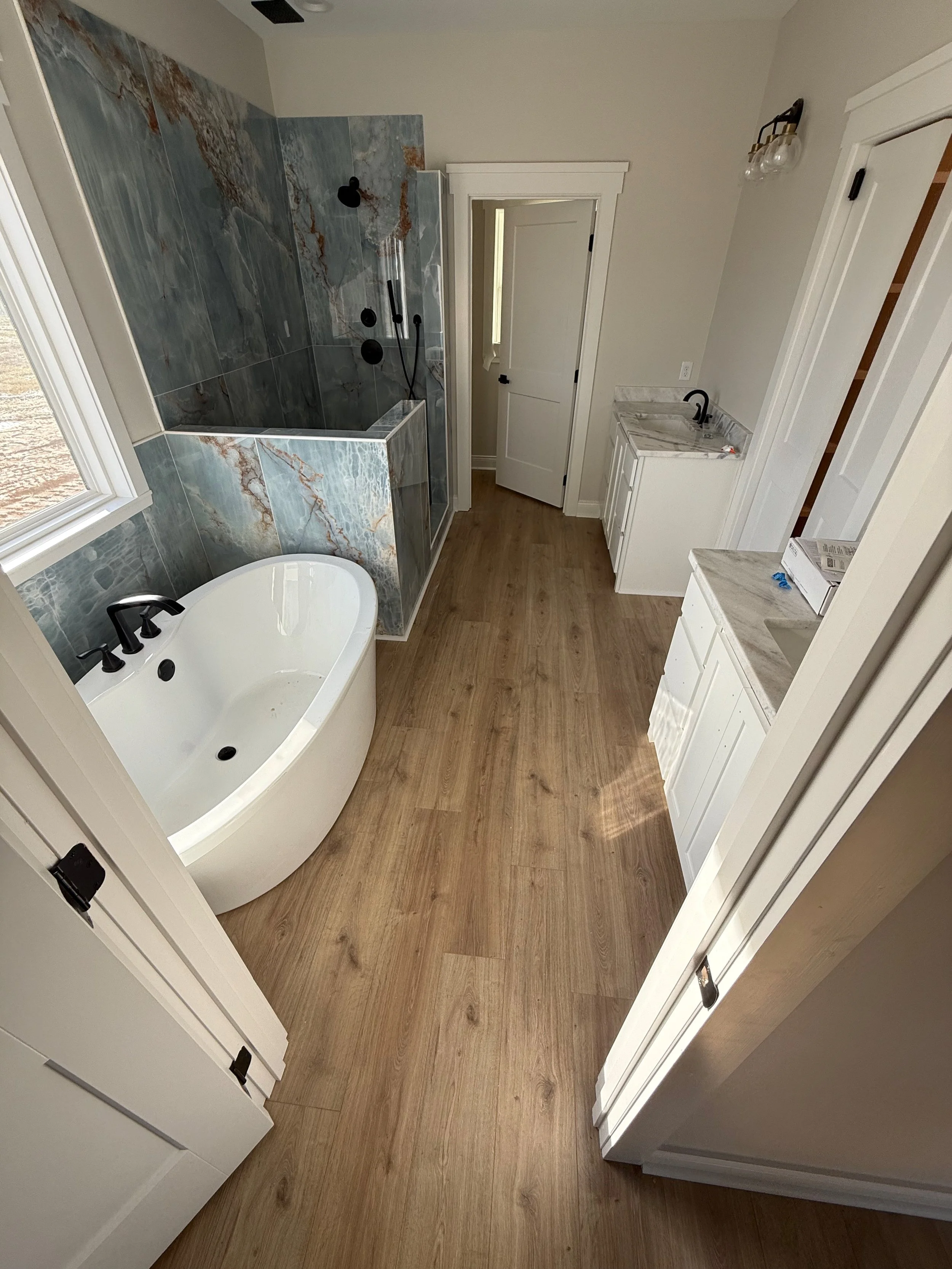A bathroom with a bathtub, a shower with blue and gray marble tiles, a white vanity with a marble countertop, a door, and wood-look flooring.