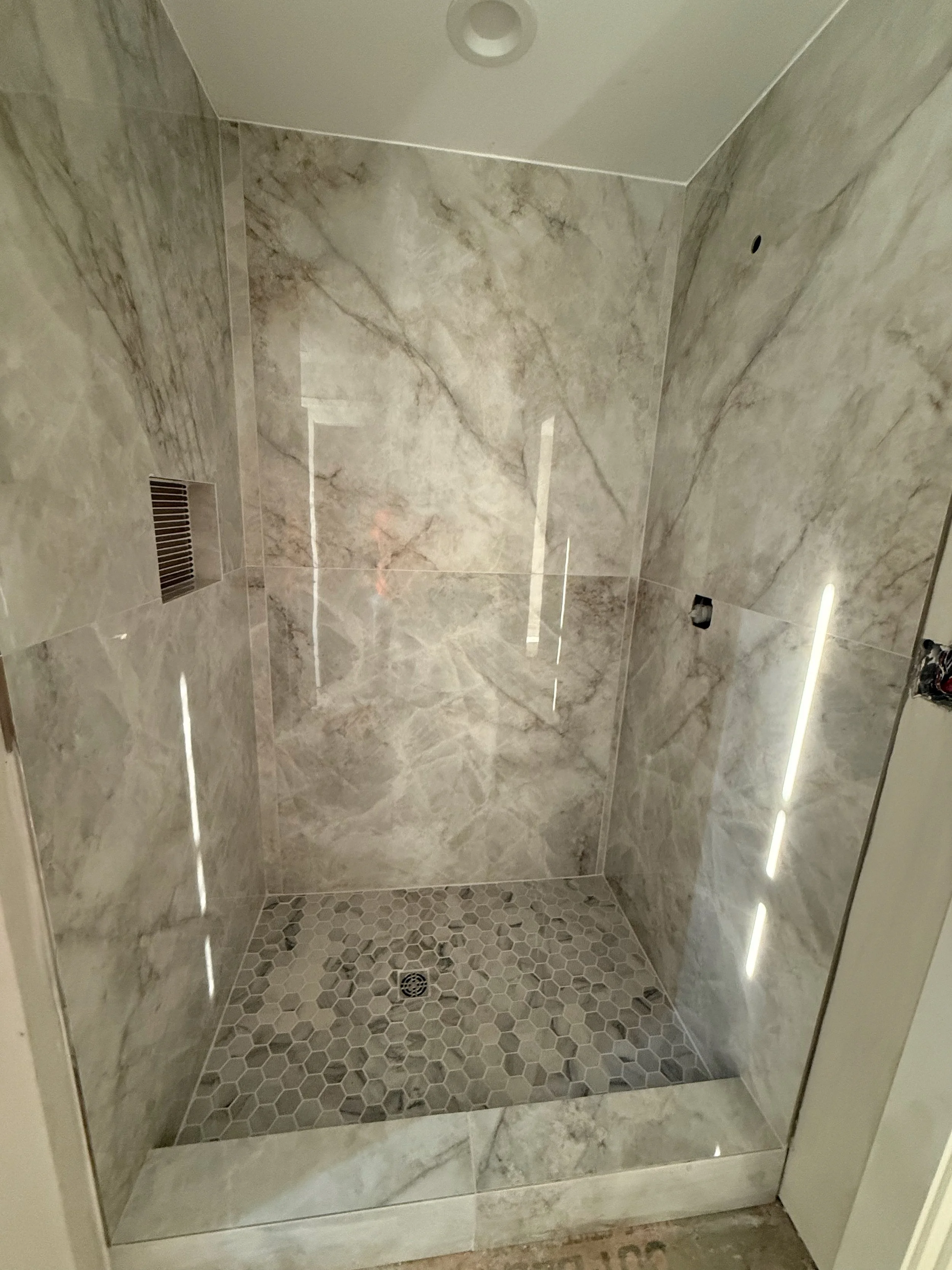 A modern shower enclosure with large marble tiles on the walls and small hexagonal tiles on the floor, a ceiling light, and a drain at the center of the floor.
