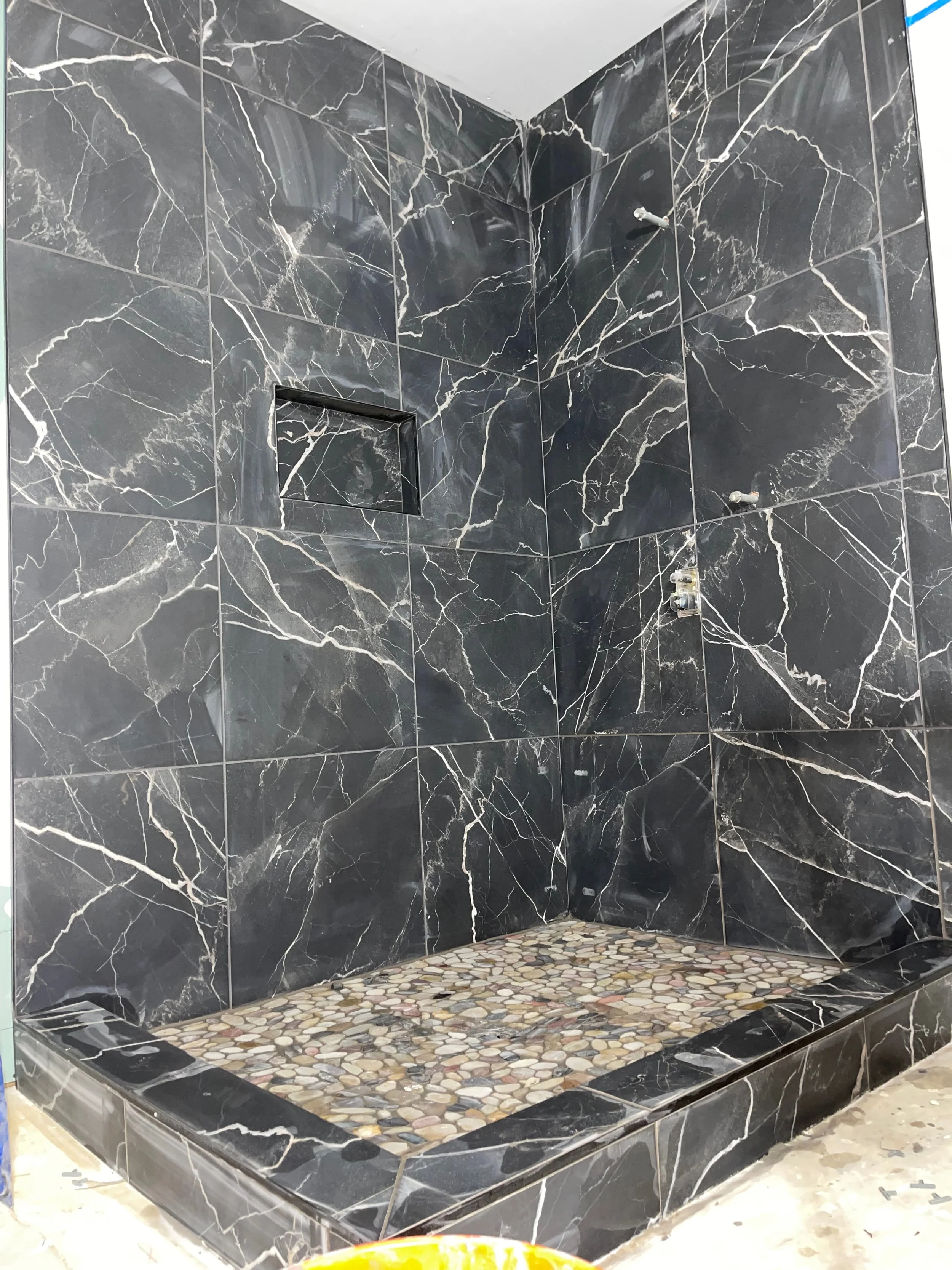 Shower area with black marble tiles, pebble floor, and built-in small shelf, no showerhead installed yet.