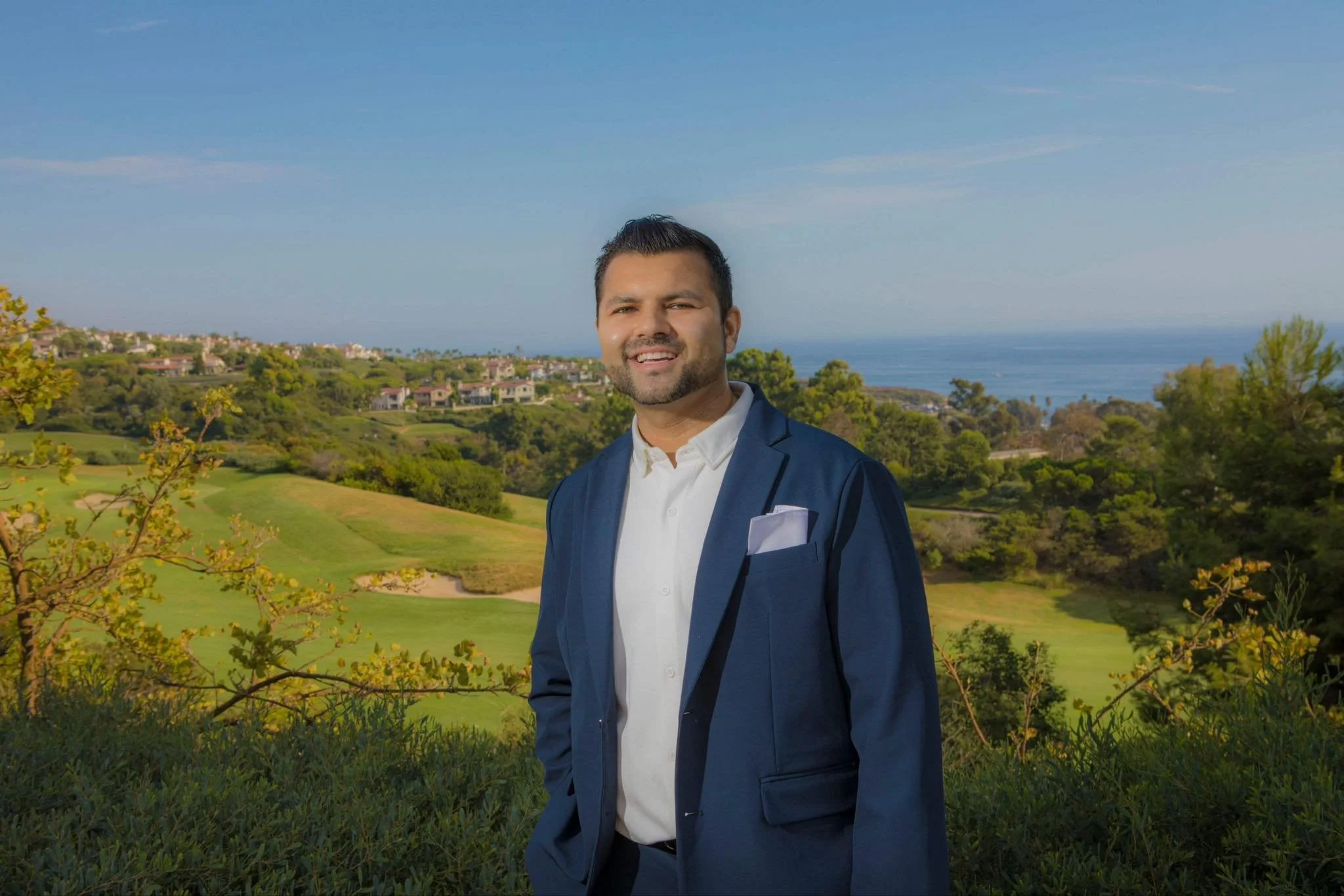 A man in a blue suit and white shirt standing outdoors with greenery, houses, and the ocean in the background, smiling at the camera.