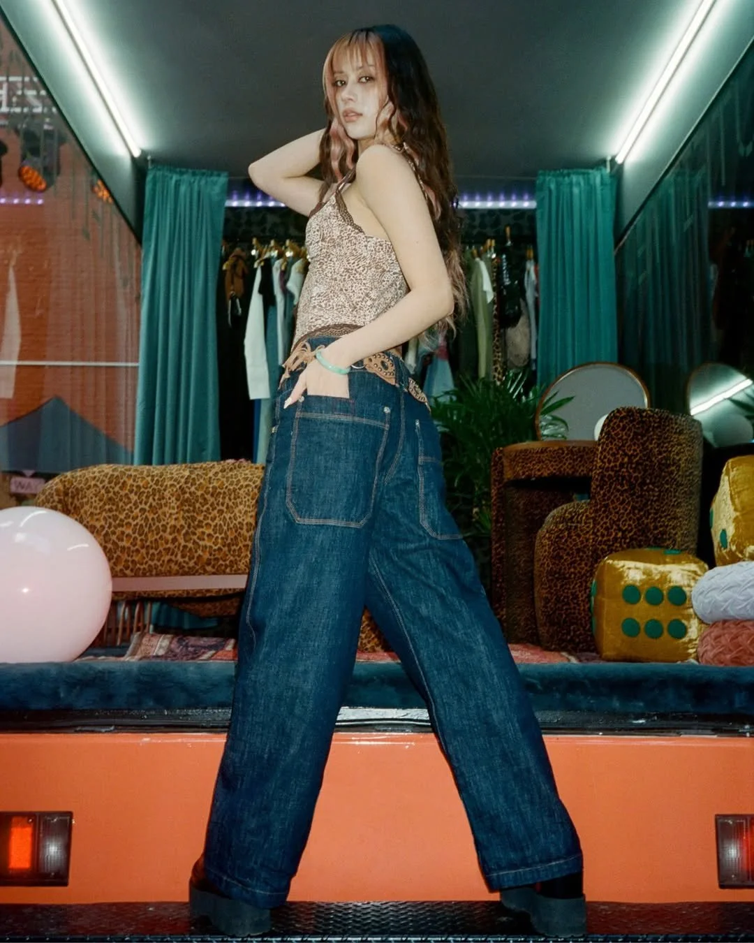 A young woman with long hair posing inside a colorful boutique or lounge area, wearing a leopard print top and baggy jeans.