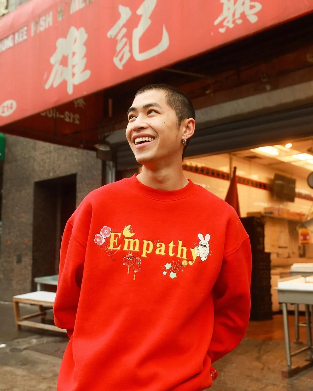 A young man with short dark hair, earrings, and a bright smile, wearing a red sweatshirt with the word 'Empathy' and colorful cartoon characters, standing outdoors in front of a restaurant with red signage.