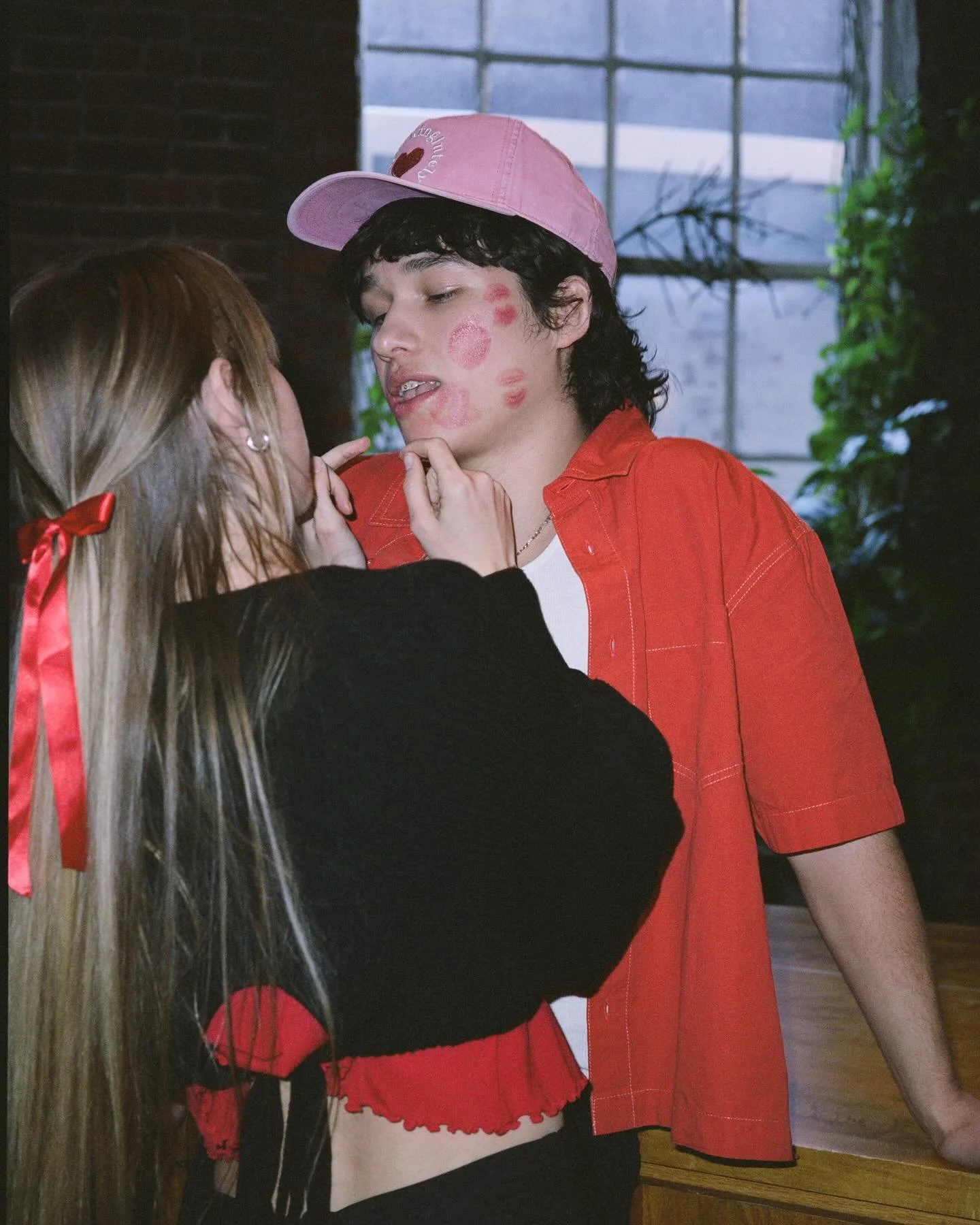 A person with long blonde hair tied with a red ribbon, wearing a black top, touches a young man's face. The young man has dark curly hair, is wearing a pink cap, a red short-sleeved shirt, and has makeup or lipstick kisses on his face.
