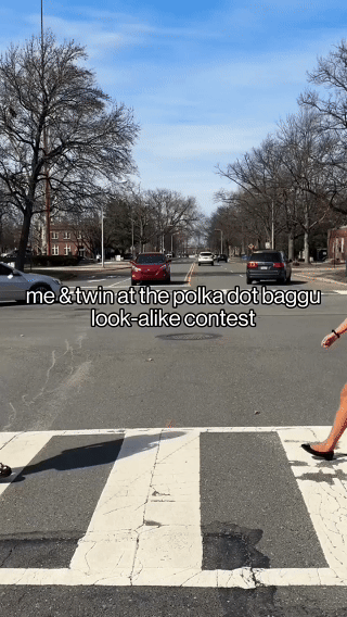 A person standing at a crosswalk with a child's arm visible on the right, watching a contest where a person and a twin dress alike at a street intersection with parked cars, leafless trees, and houses in the background.