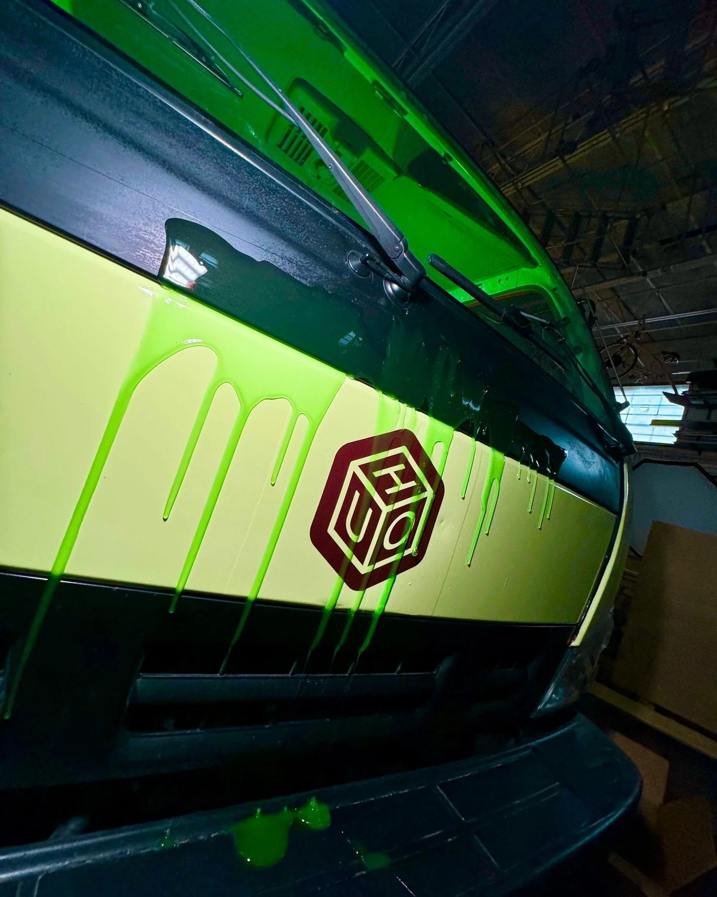 A green and yellow vehicle with a black windshield wiper and a red logo on its side. The background shows a ceiling with indiscernible structures.