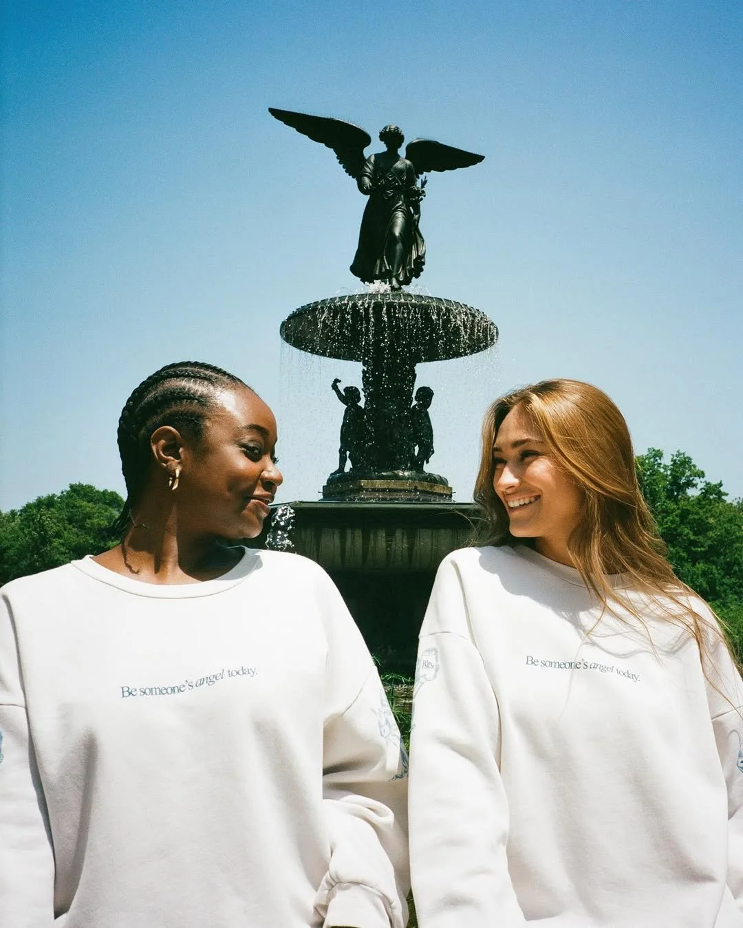 Two women standing in front of a fountain with a statue of an angel on top, both wearing white sweatshirts that say 'Be someone's angel today', smiling at each other.