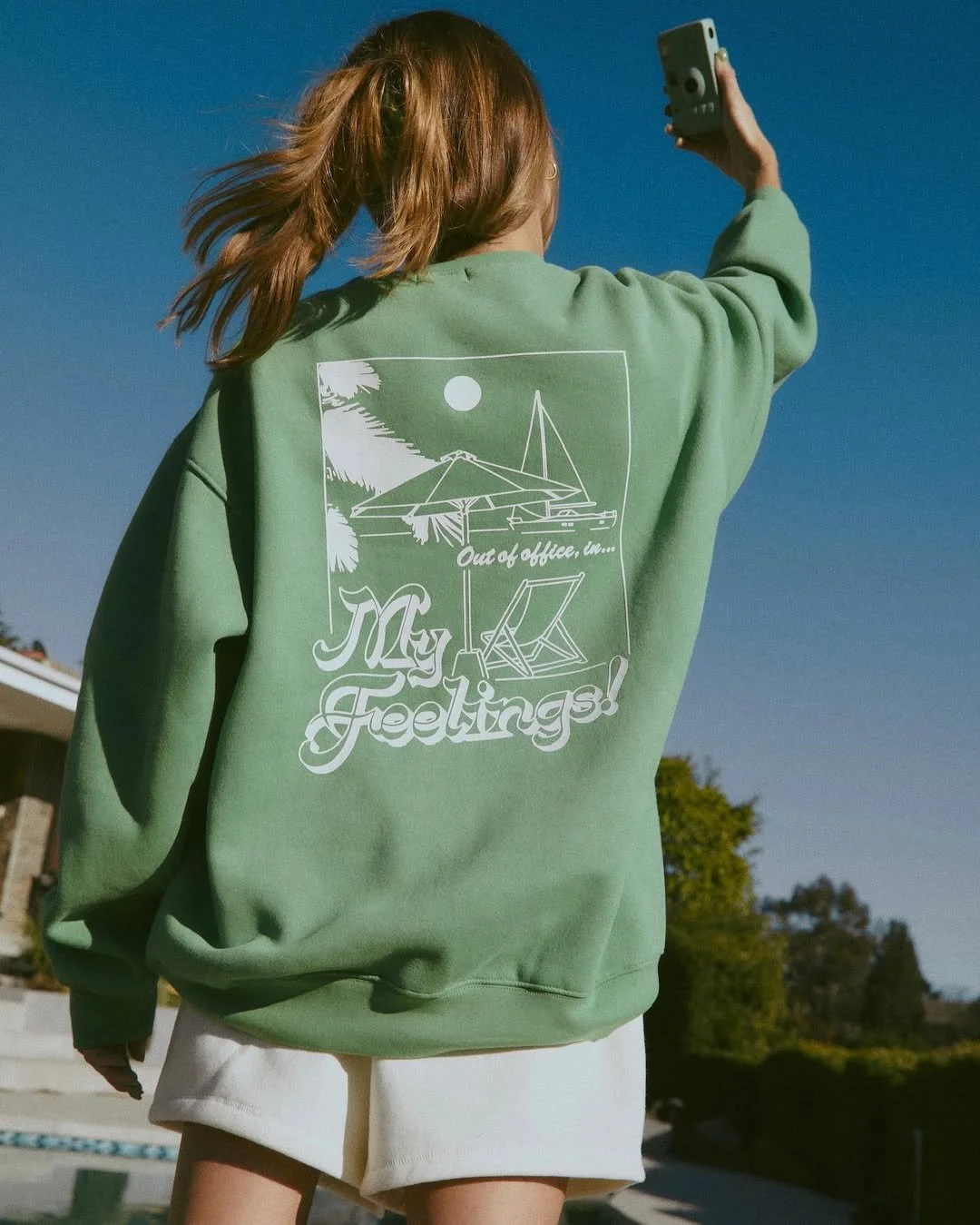 Back of a woman taking a selfie, wearing a green sweatshirt with a beach scene, text reading "My Feelings!" and "Out of office, here" printed on it, outdoors with blue sky and trees in the background.