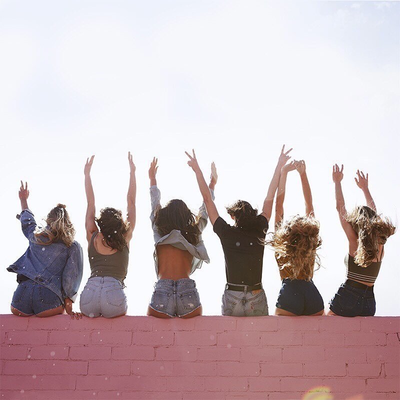 Six young women sitting on a pink wall with their backs to the camera, raising their arms in the air during daylight.