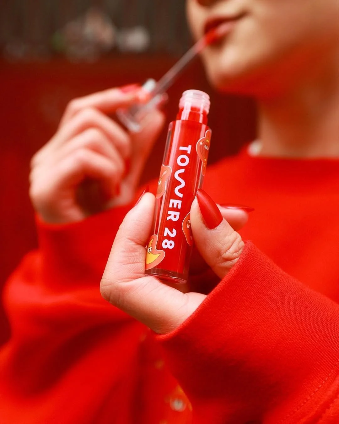 Close-up of a hand with red-painted nails holding a red lip gloss tube labeled 'TOWER 28' with a small duck illustration, while the person applies lip gloss with a wand.