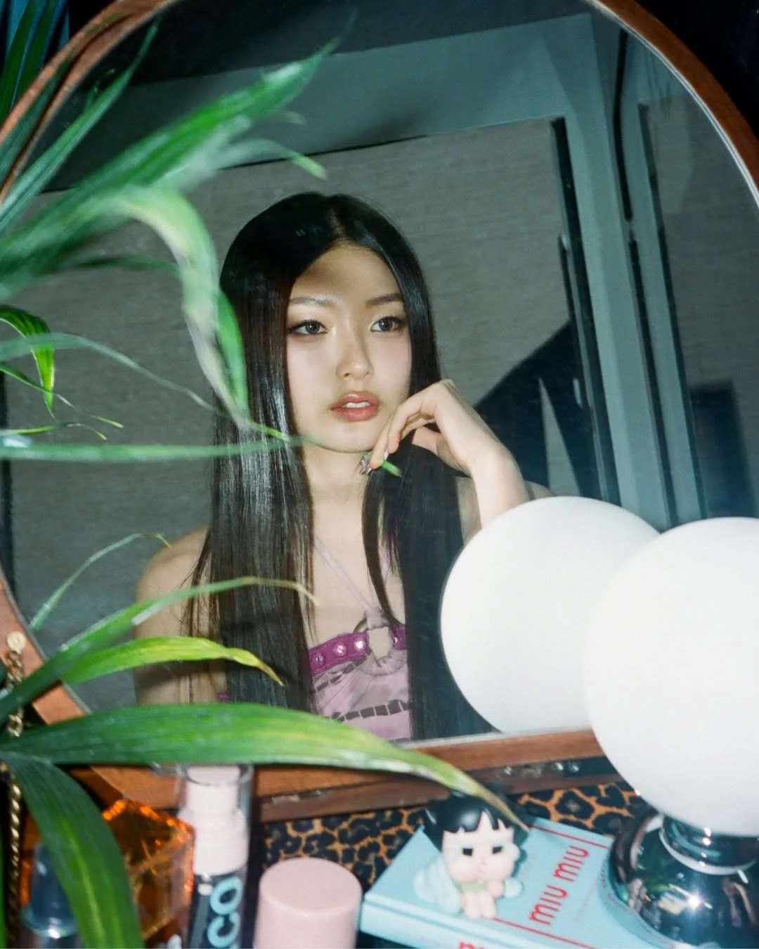 A young woman with long black hair looks through a mirror, touching her face with her hand, with makeup and a pink top visible. A large mirror and various personal items are on a vanity table in front of her.