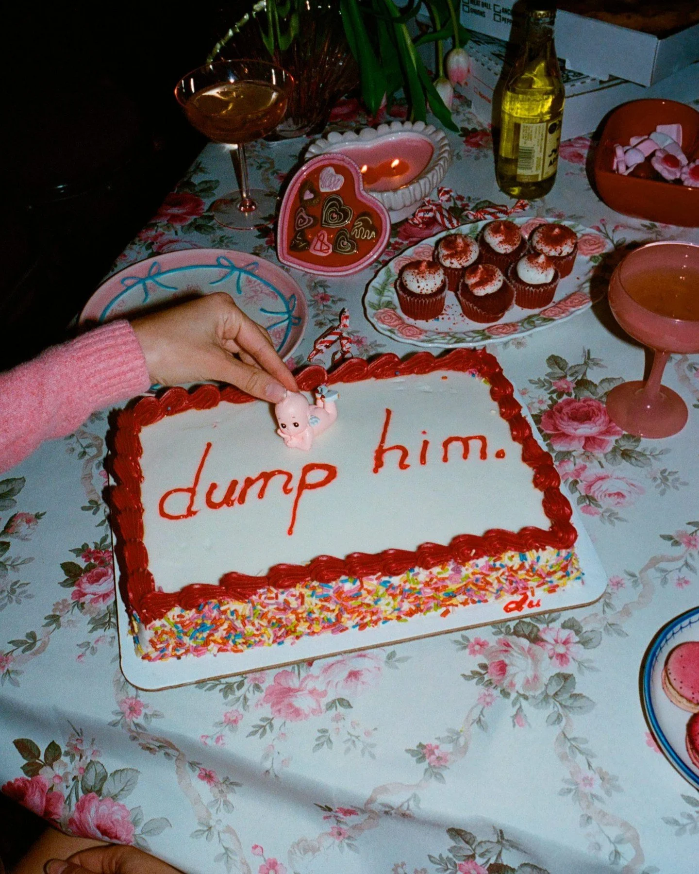A birthday celebration table with a cake that has the words 'dump him' written on it in red icing, surrounded by cupcakes with white frosting and red sprinkles, balloons, drinks, and a Valentine's Day themed heart decoration.