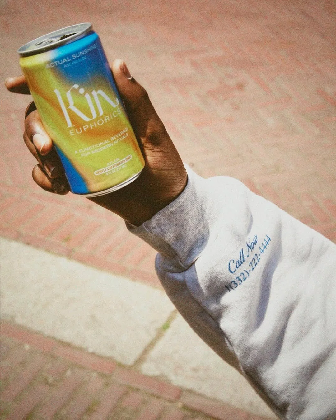 Hand holding a colorful can of Kin Euphoric beverage outdoors, with brick sidewalk in the background.
