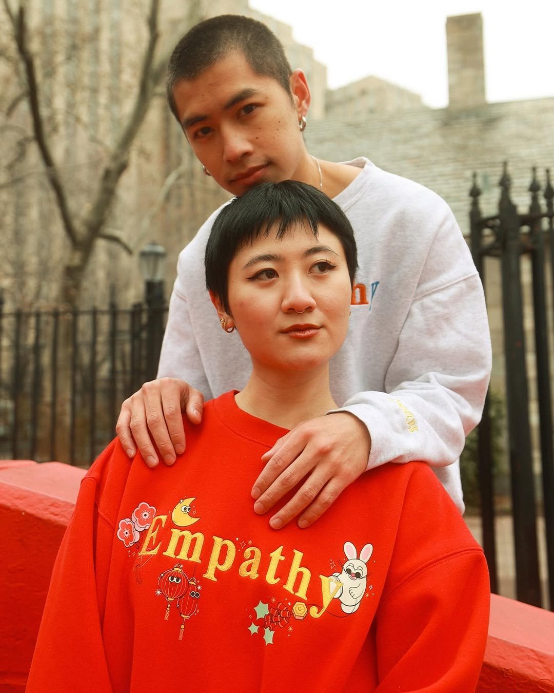 A man and woman posing outdoors. The man stands behind the woman with his hand on her shoulder; he has short hair, earrings, and a light gray sweatshirt. The woman has short dark hair and is wearing a red sweatshirt with colorful graphics and the word 'Empathy' on it. They are in front of a black metal fence and fall trees, with a cloudy sky in the background.