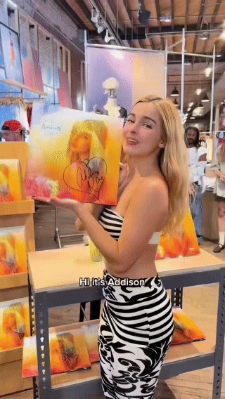 A young woman with blonde hair wearing a black and white striped top and patterned skirt holds a colorful art print at an indoor art store. She is smiling and standing next to a display of similar prints, with the caption "Hi it's Addison". The backg