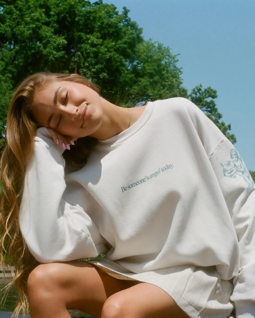 A young woman with long, wavy hair is sitting outdoors with her eyes closed, leaning her head on her hand and smiling gently. She is wearing a white sweatshirt with a small text and illustration on it, and green shorts. The background features green trees and a clear blue sky.