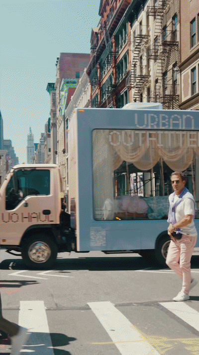 A city street scene with a person crossing a crosswalk, a delivery truck, and a small mobile structure with the words 'URBAN' and 'OUTFITTERS' visible.