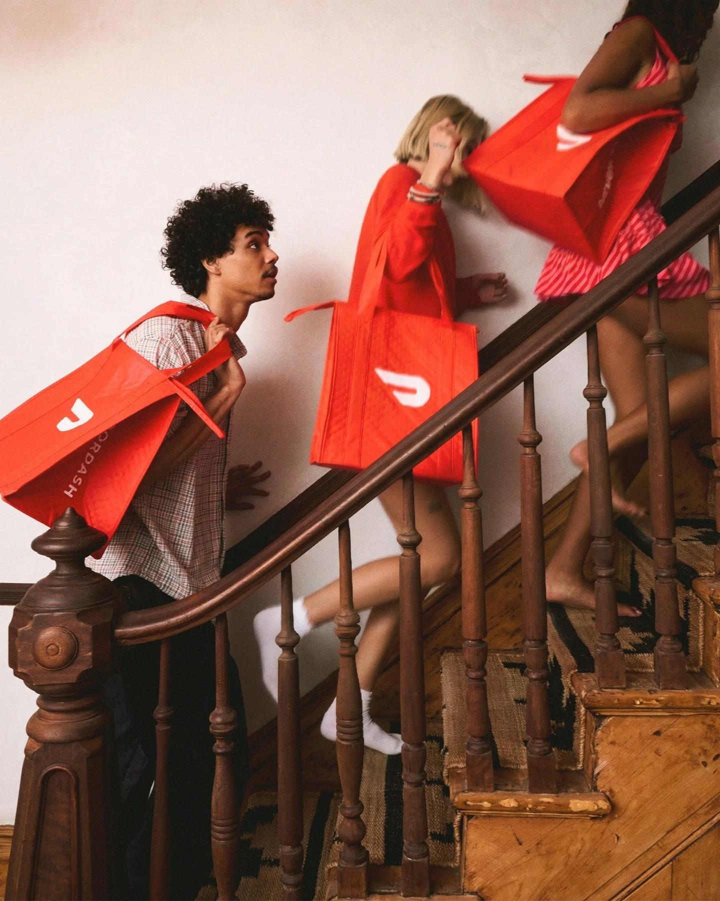 Three women, two standing and one lying on the staircase, all wearing red tote bags and pajamas, engaged in a playful scene on a wooden staircase inside a home.