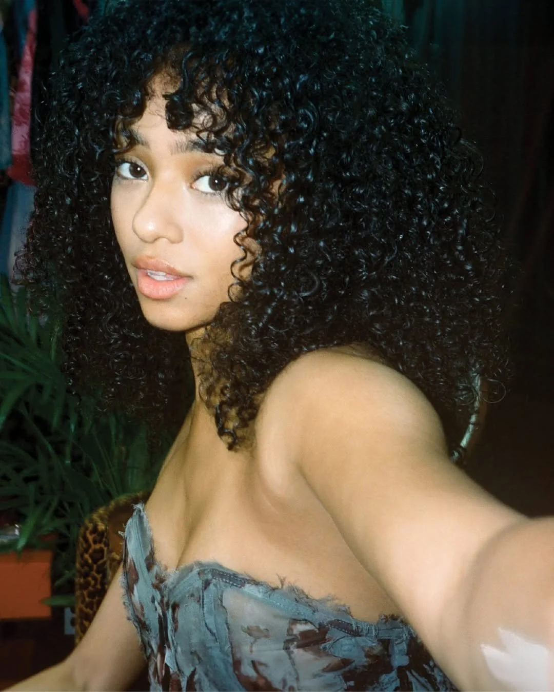 A young woman with medium skin tone and dark, curly hair taking a selfie indoors, with a slight smile and wearing a sleeveless top.