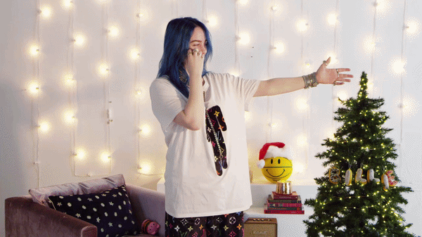 A woman with blue hair standing in a decorated room with Christmas lights, a small Christmas tree, and holiday decor, talking on her phone.