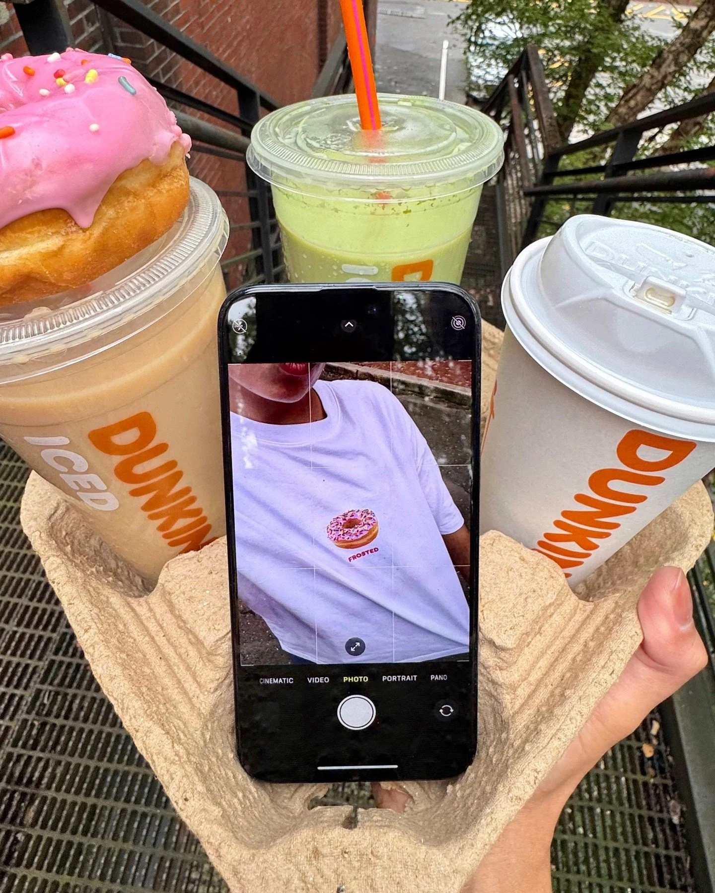 Close-up of a person's hand holding a cardboard tray with three Dunkin' cold beverages and a donut with pink icing and sprinkles, with a smartphone capturing the scene. The drinks include a pink iced coffee, a green smoothie with a straw, and a white